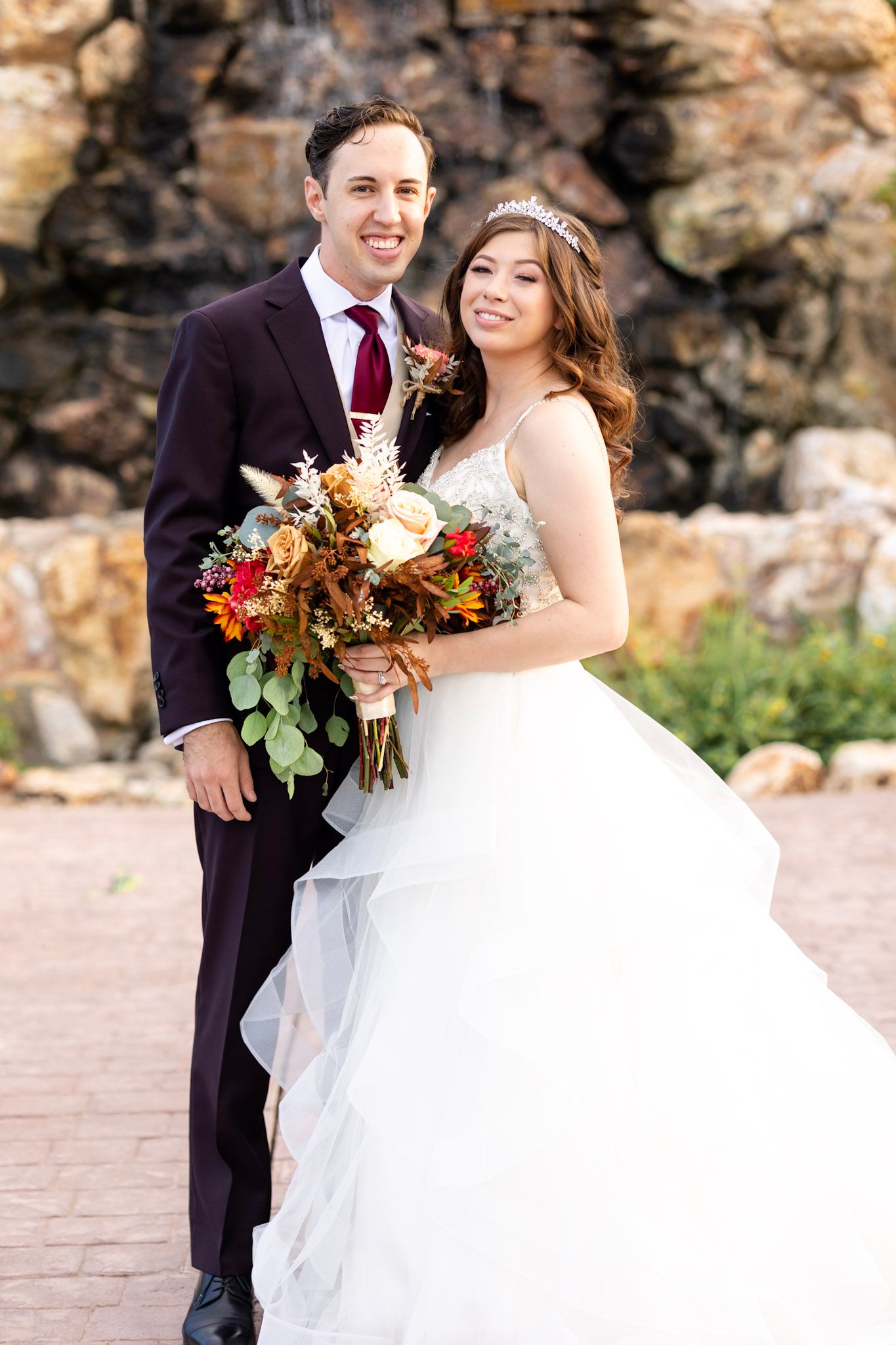 Elegant fall wedding couple poses against stone wall backdrop with autumn-colored bouquet.