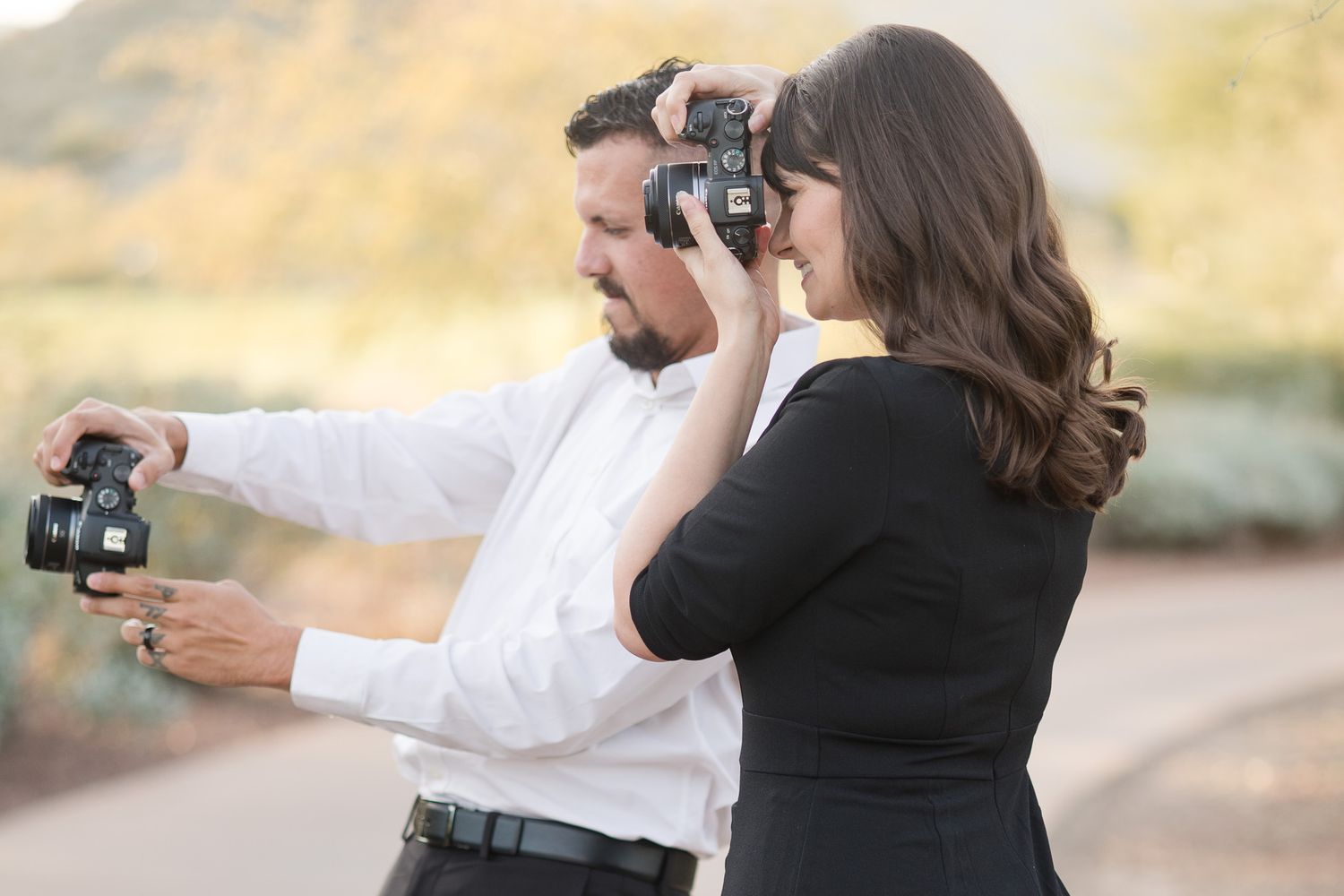 Two photographers stand side by side outdoors taking pictures with cameras in warm natural lighting.