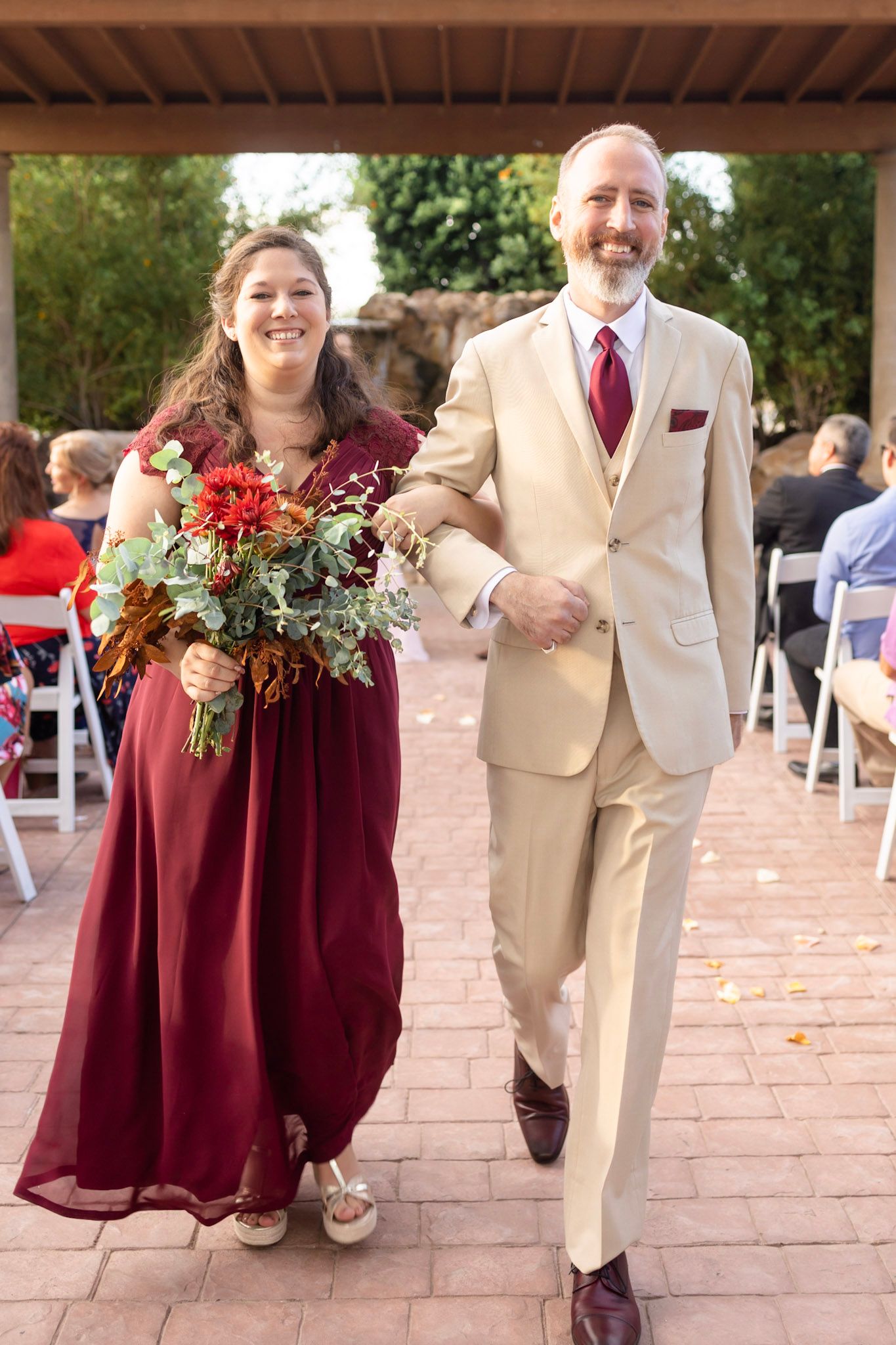 Wedding attendants in burgundy dresses and tan suits walk arm-in-arm down a brick courtyard.