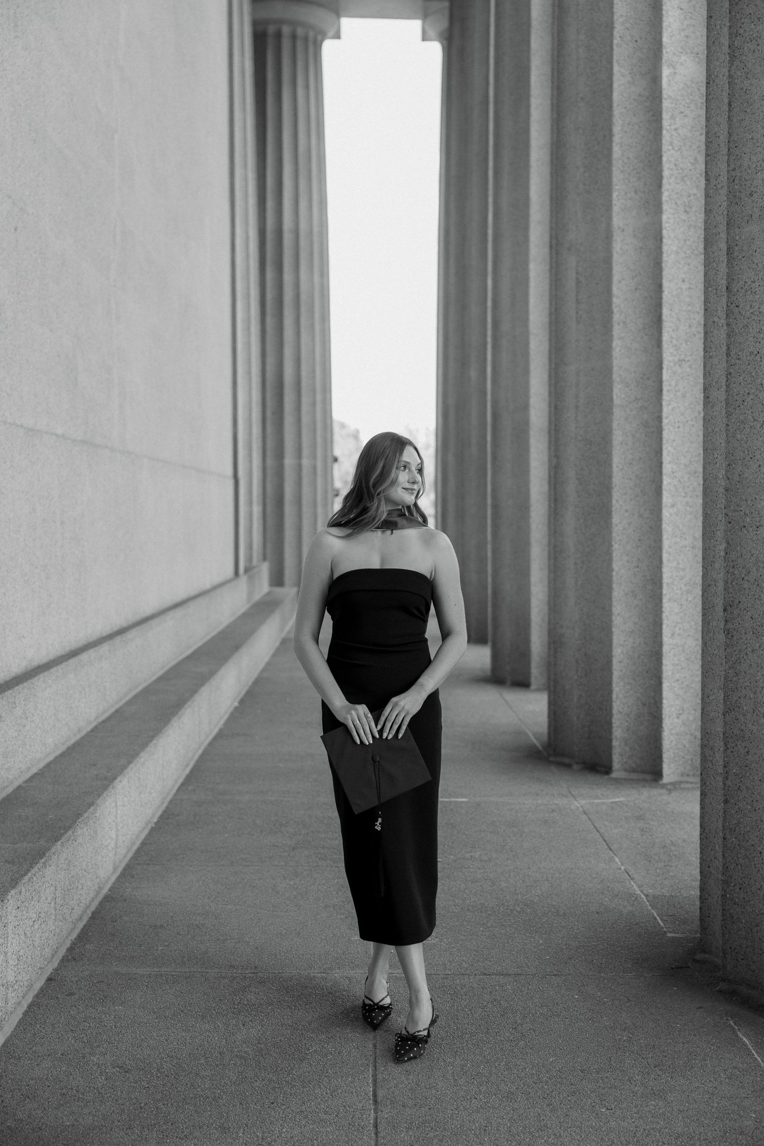 Woman in black strapless midi dress holding clutch, posing between grand stone columns in black and white photo.