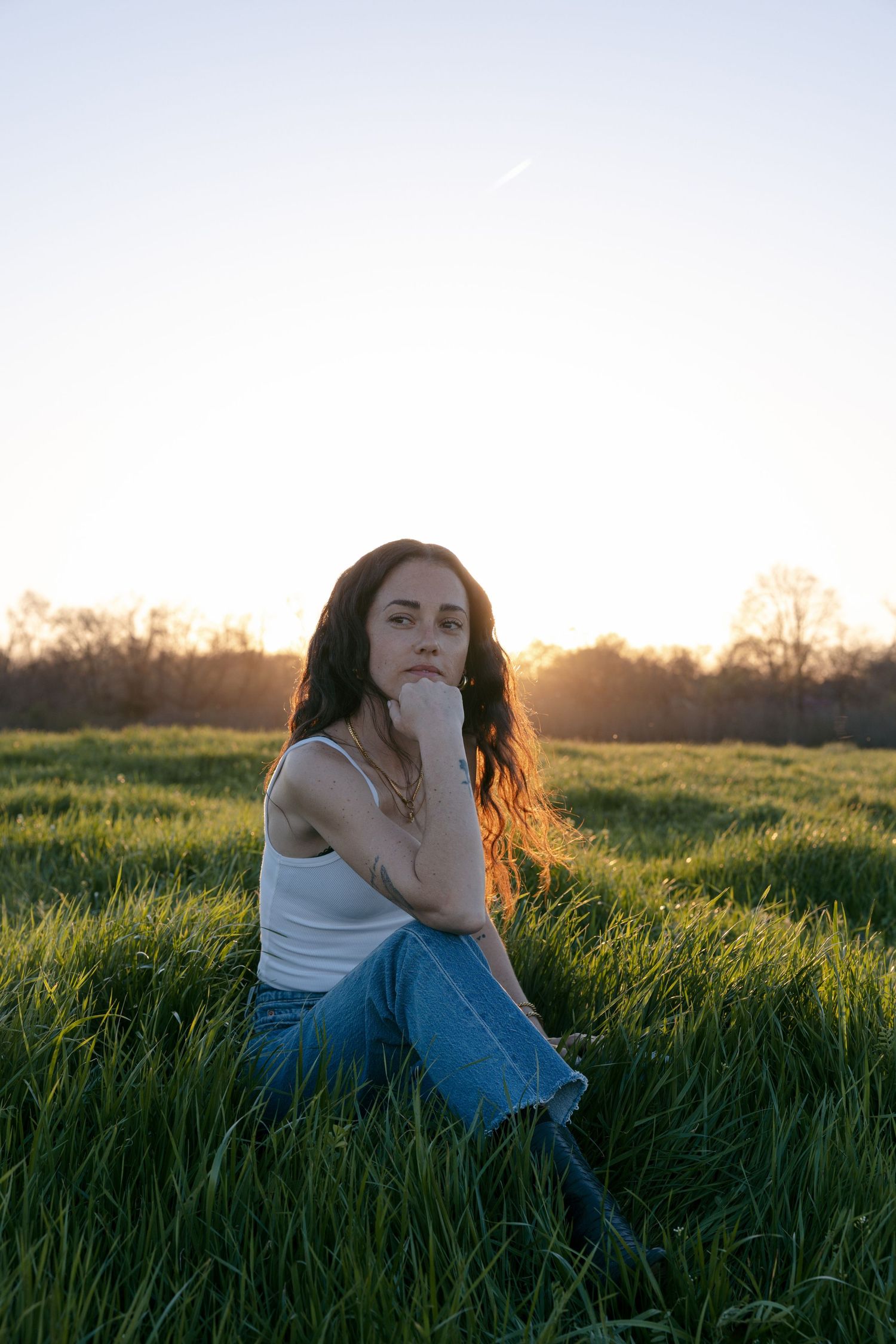 Person sitting in a grassy field at sunset wearing a white tank top and jeans while posing thoughtfully.