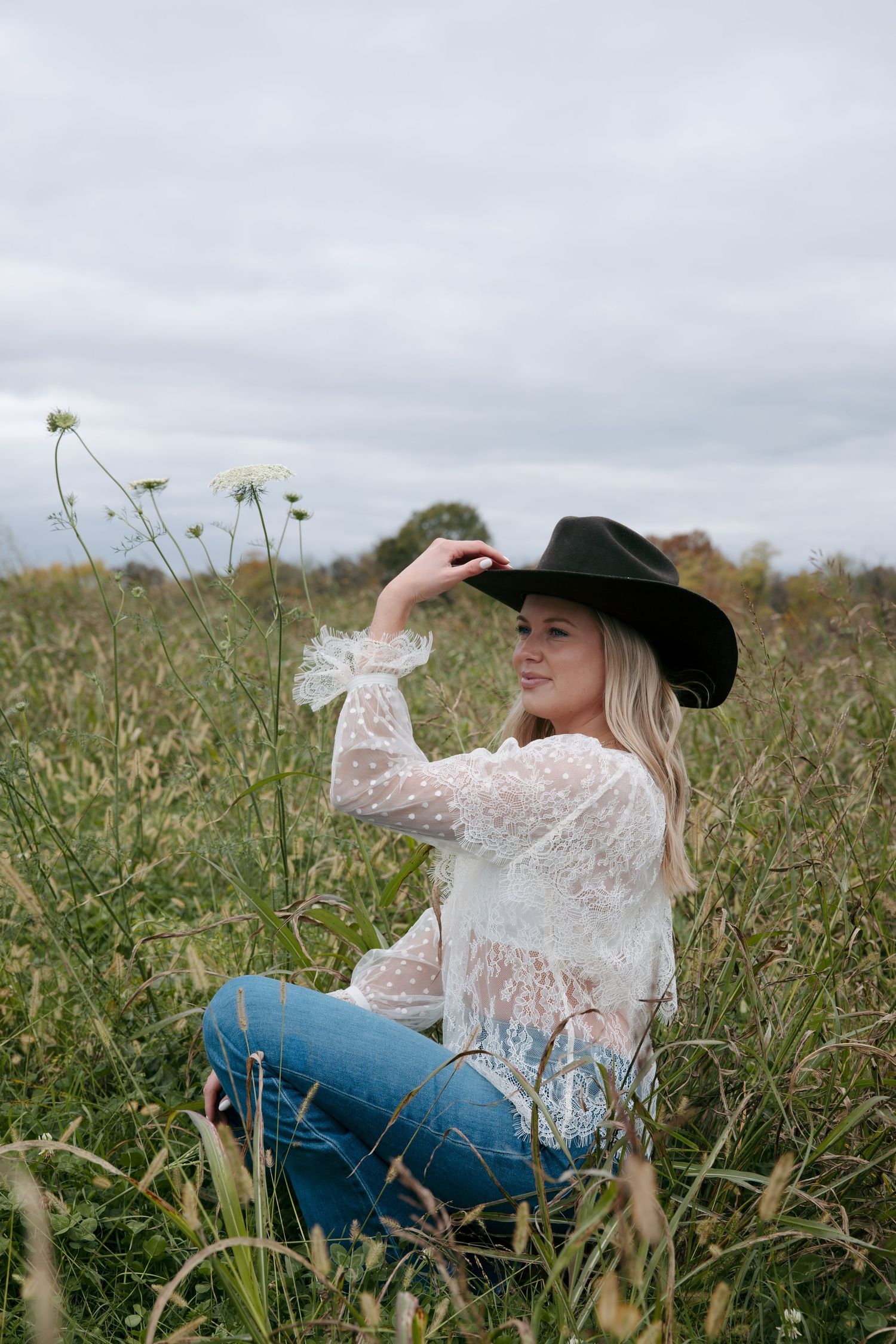 A person in a lace top and jeans lounges in a field of tall grass on an overcast day.