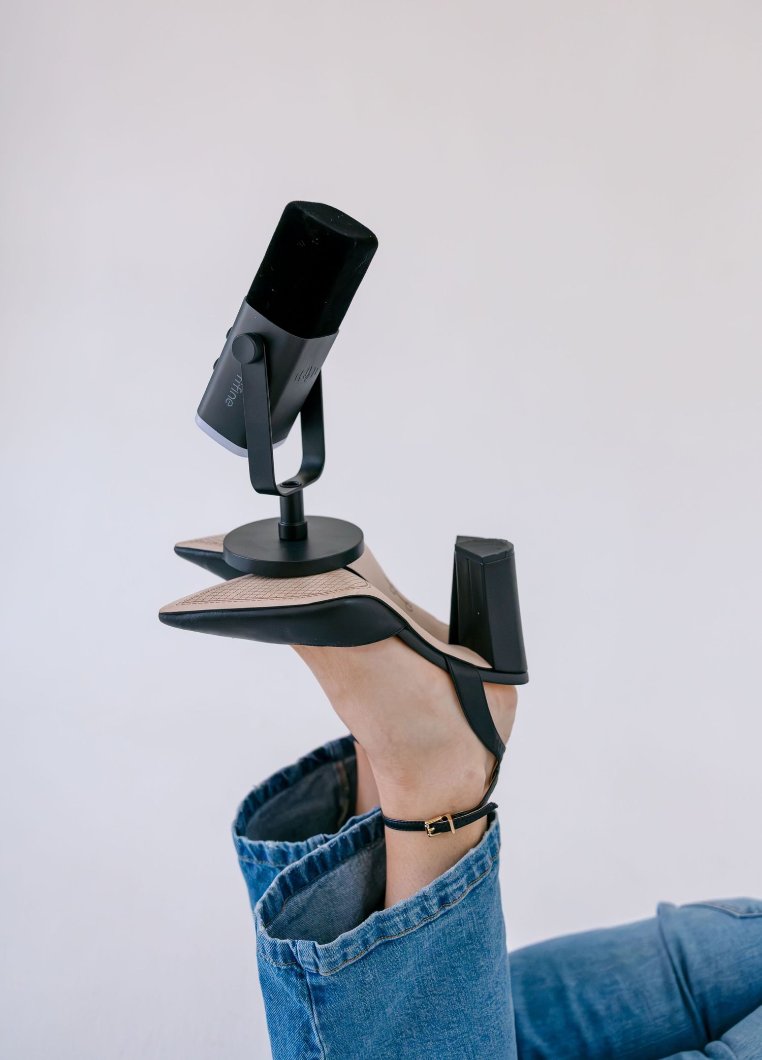 A person balancing a black microphone on their heel while wearing beige stilettos and blue jeans against a white background.