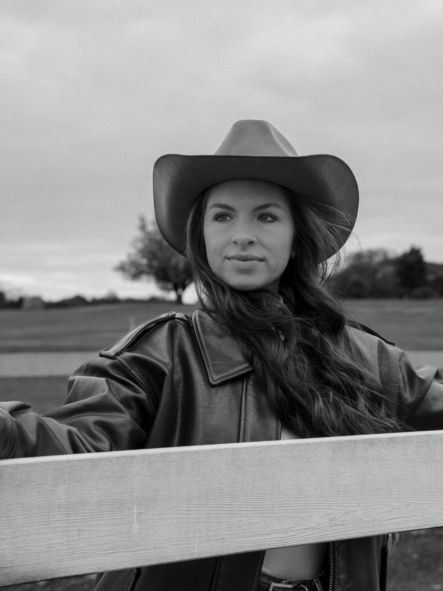 Black and white panoramic image shows someone in a cowboy hat leaning against a wooden fence in a rural setting.