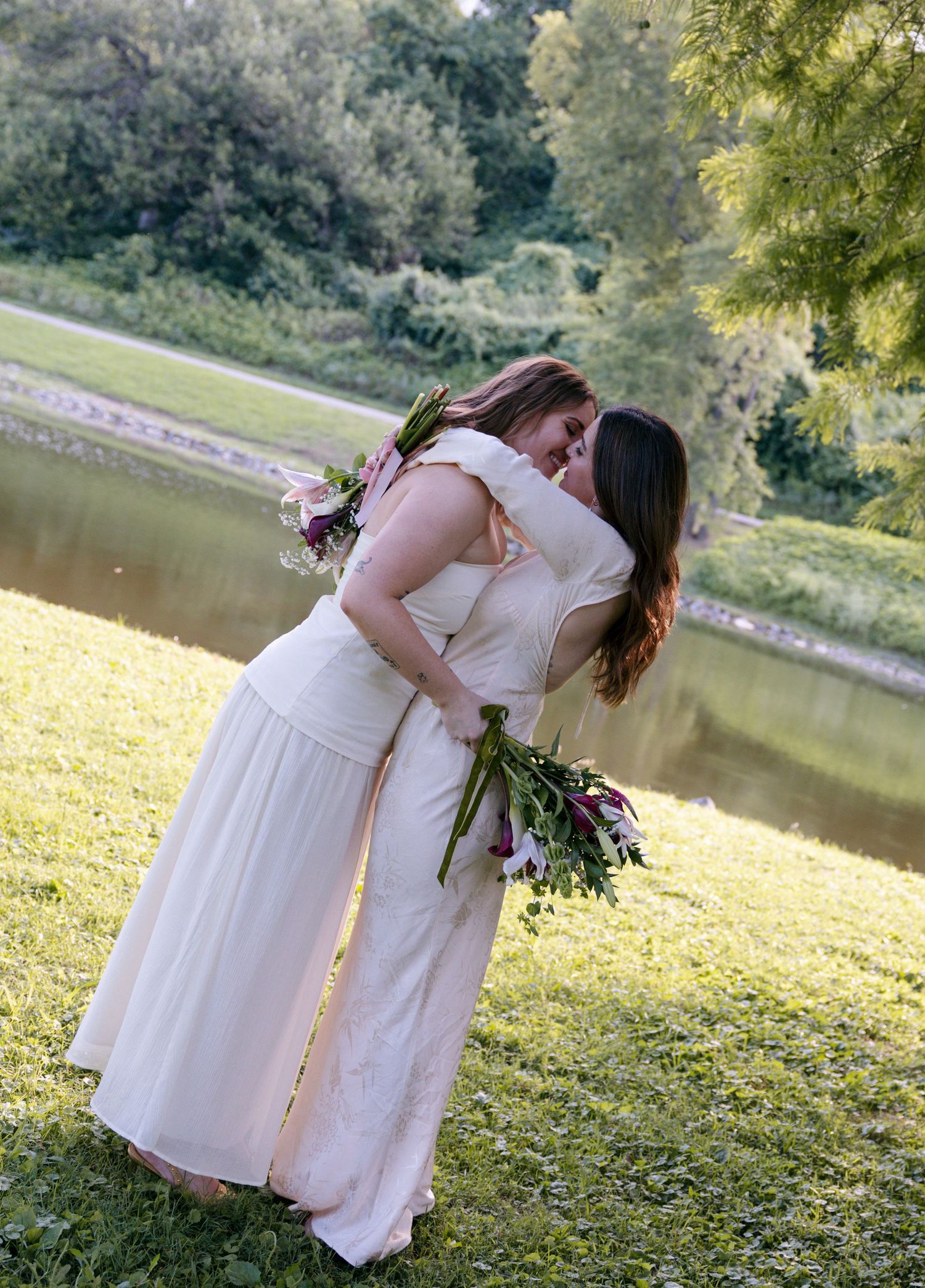 Two brides in white dresses share a kiss while holding purple floral bouquets outdoors near a pond.