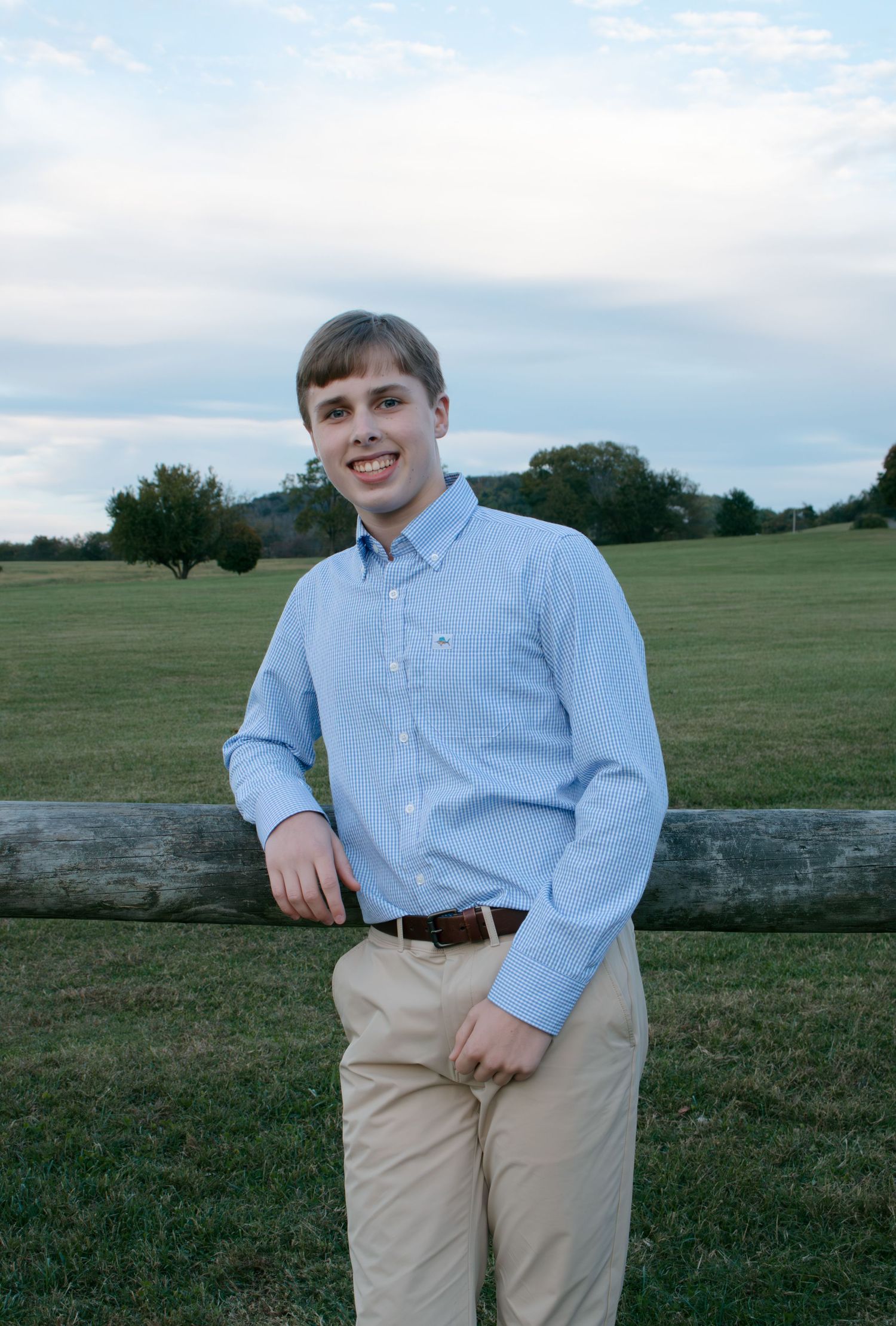 Someone wearing a light blue button-down shirt and khaki pants stands by a wooden fence in a grassy field on a cloudy day.