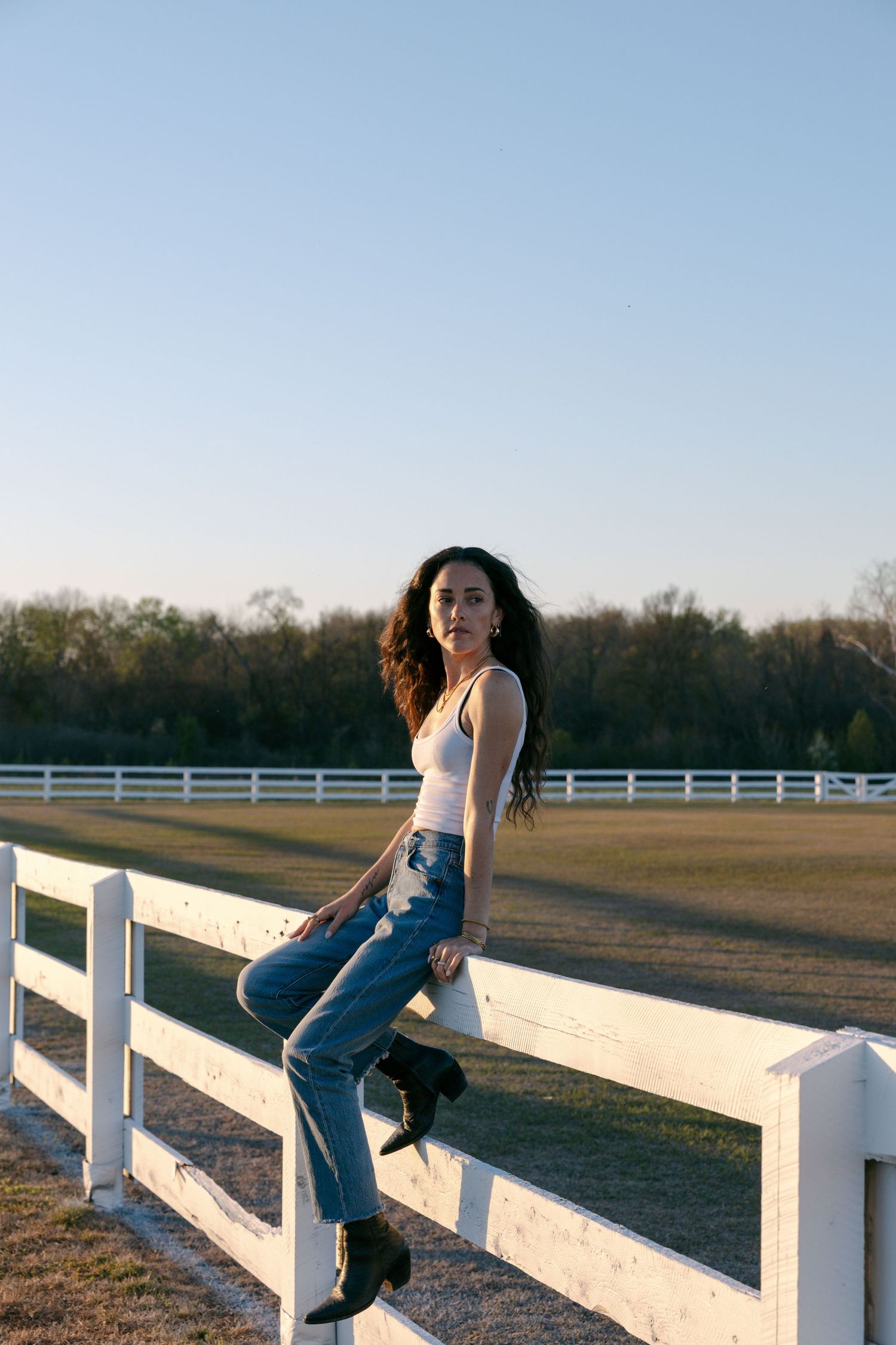 Person in casual denim sits on a white wooden fence at sunset with rustic farmland in the background.