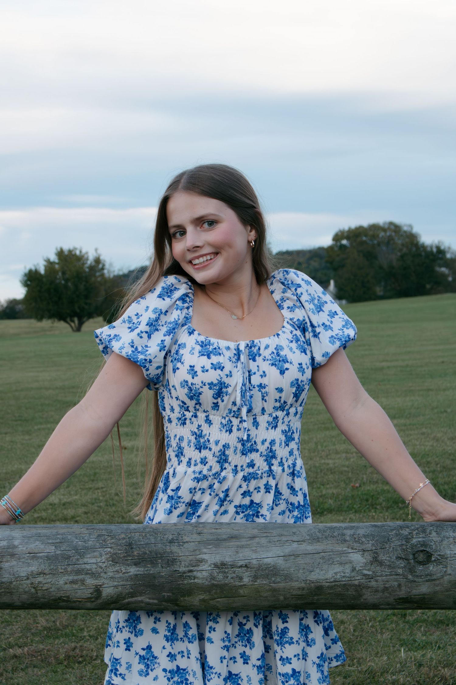 A person in a blue and white floral dress leans against a rustic wooden fence in a pastoral countryside setting.