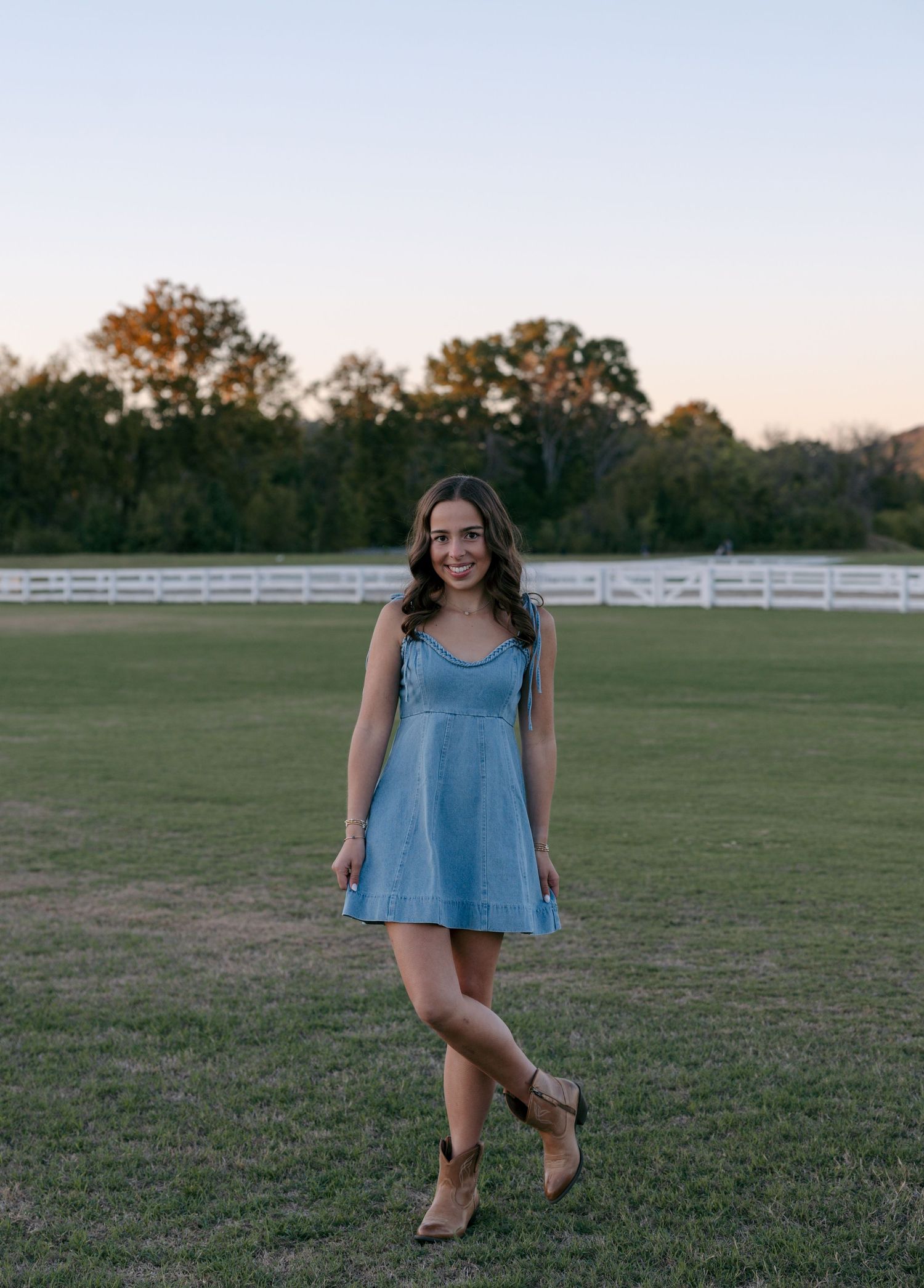 Smiling woman in a short blue denim dress and tan cowboy boots stands on a grassy field with a white fence and trees at sunset.