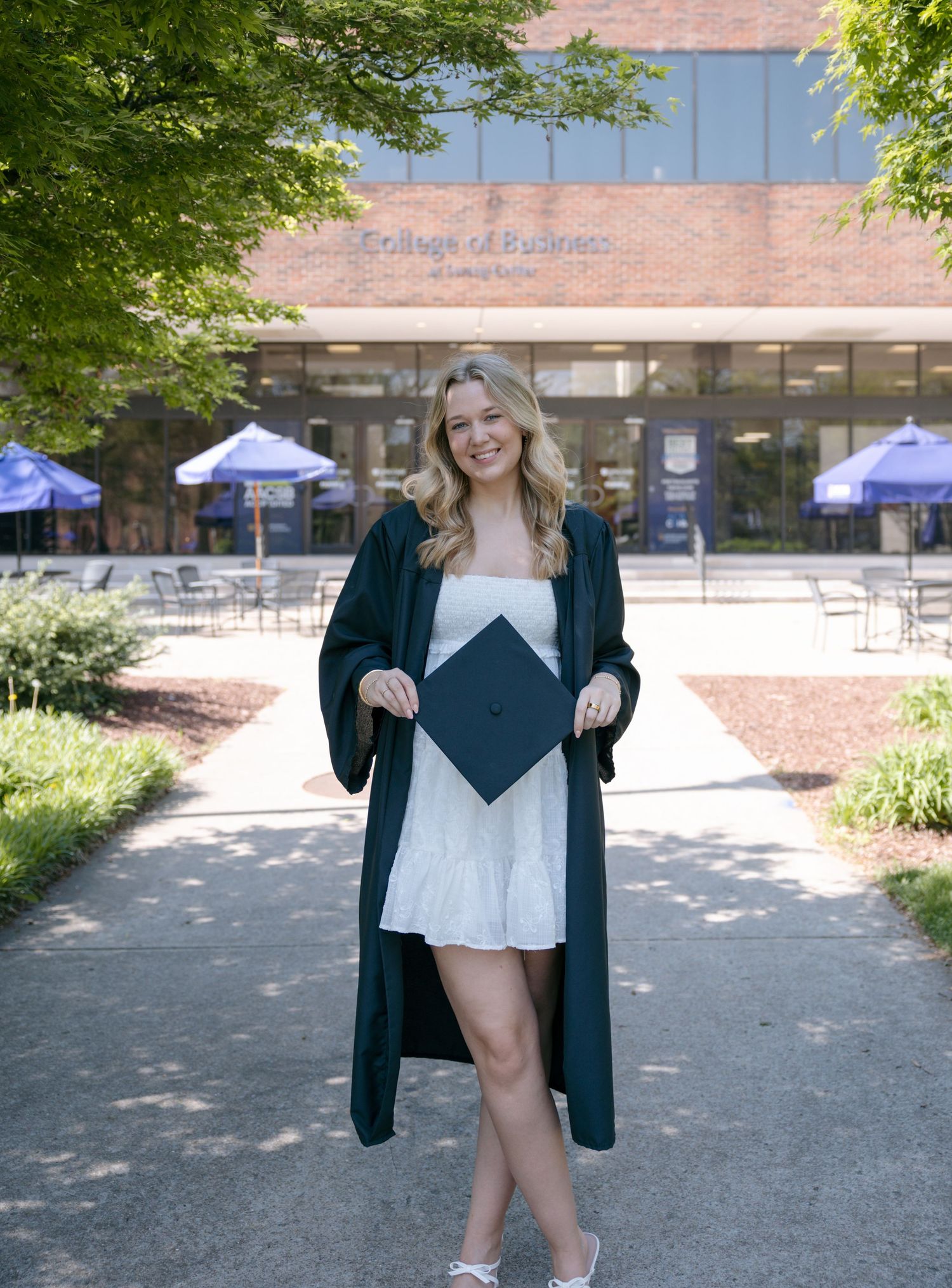 Smiling graduate in green robe holds cap outside College of Business building on sunny campus.
