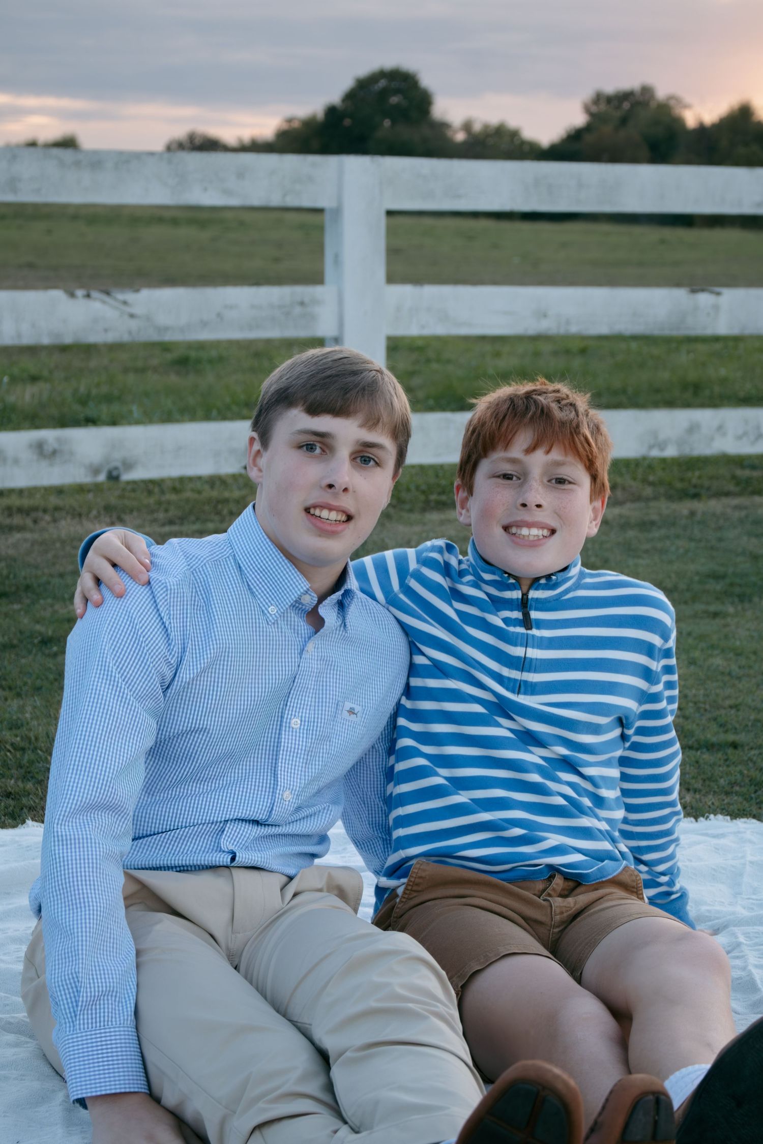 Two children sit together on a blanket outdoors wearing coordinating blue and khaki outfits at sunset.