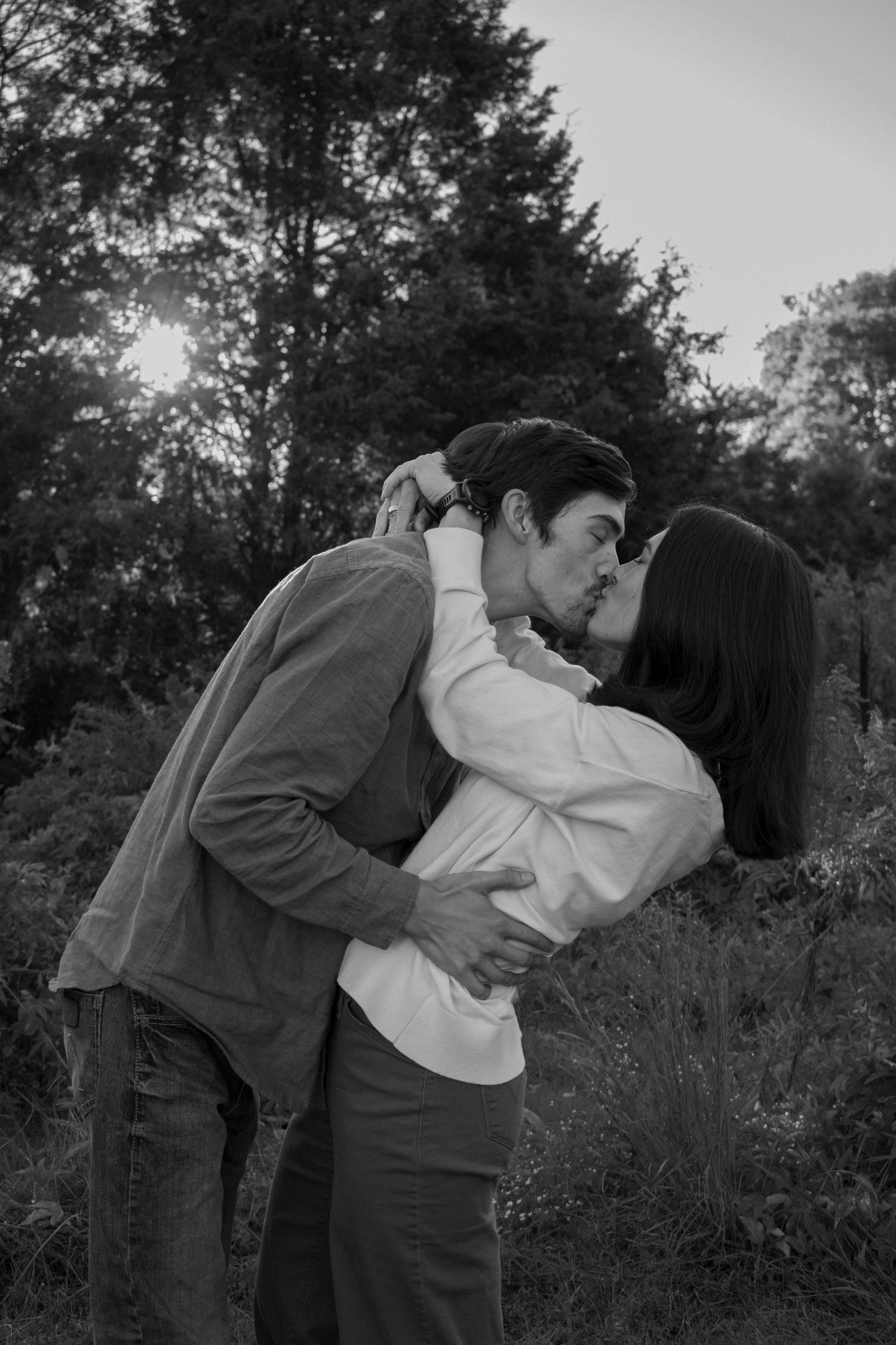 A romantic black and white photo of a couple kissing in a wooded setting at sunset.