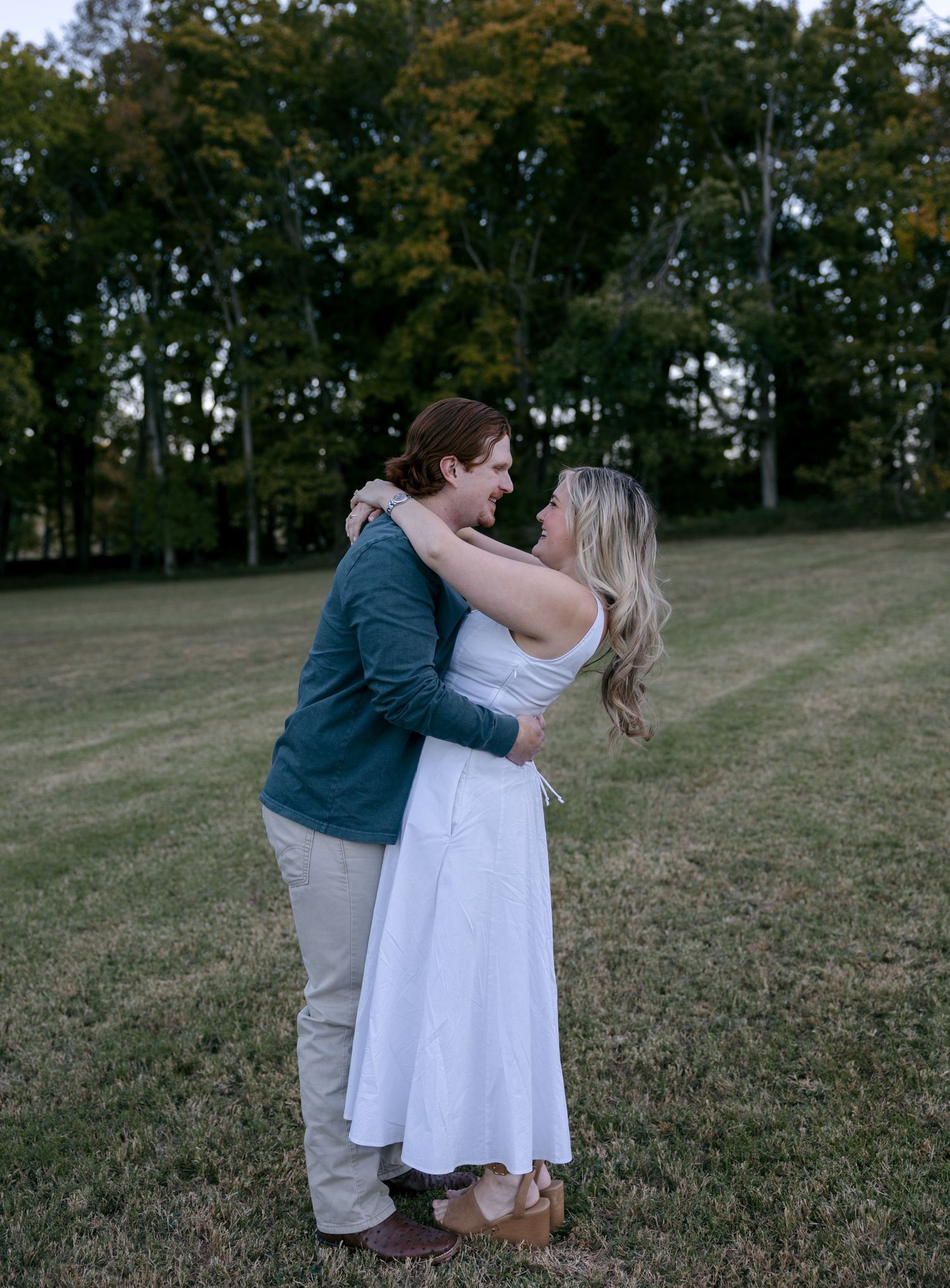 Couple embracing in a grassy field at dusk, the woman in a white dress and the man in a teal shirt, with trees behind them.