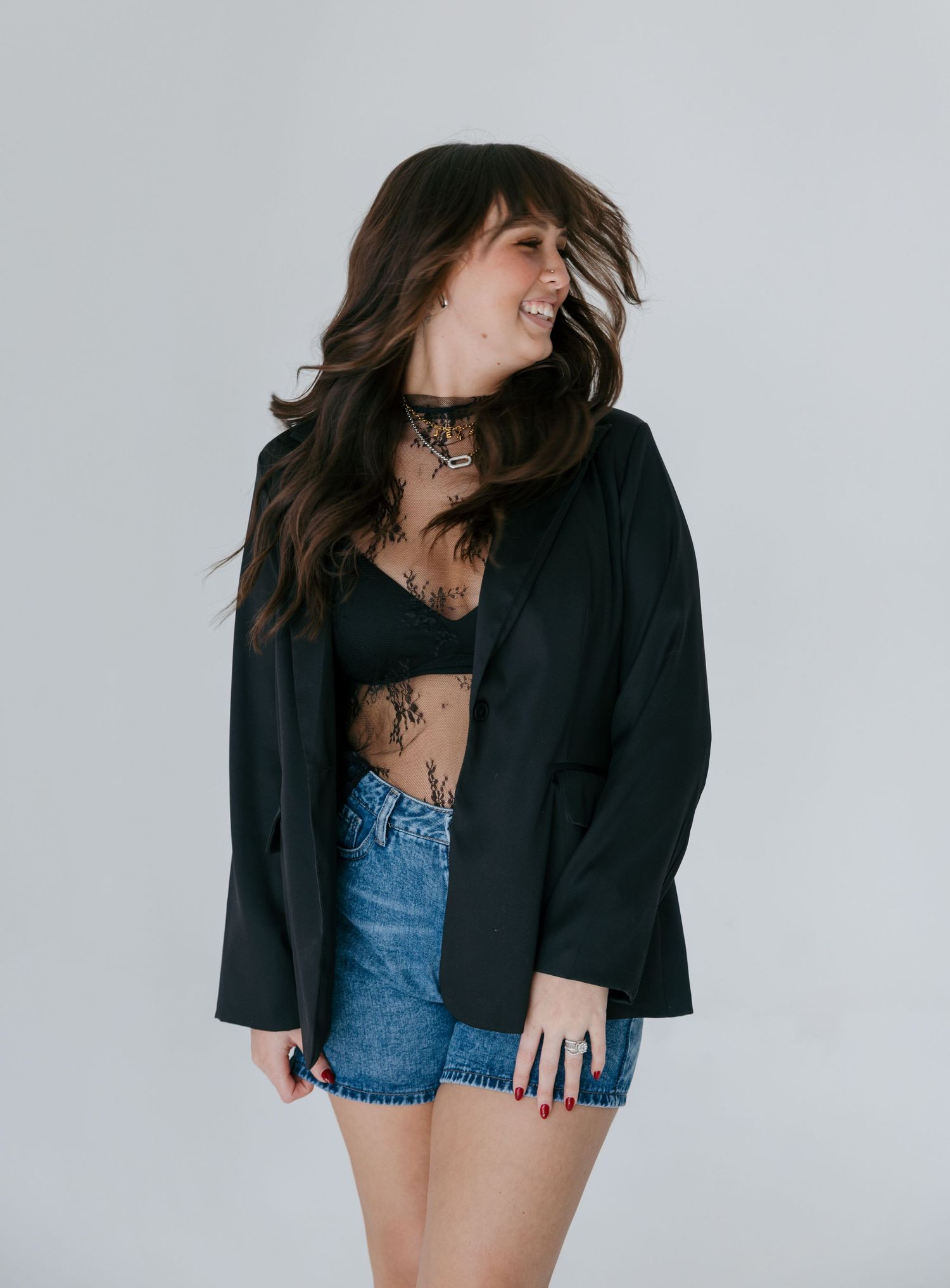 Woman in black blazer, crop top, and denim shorts smiling, with long dark wavy hair and a choker necklace.