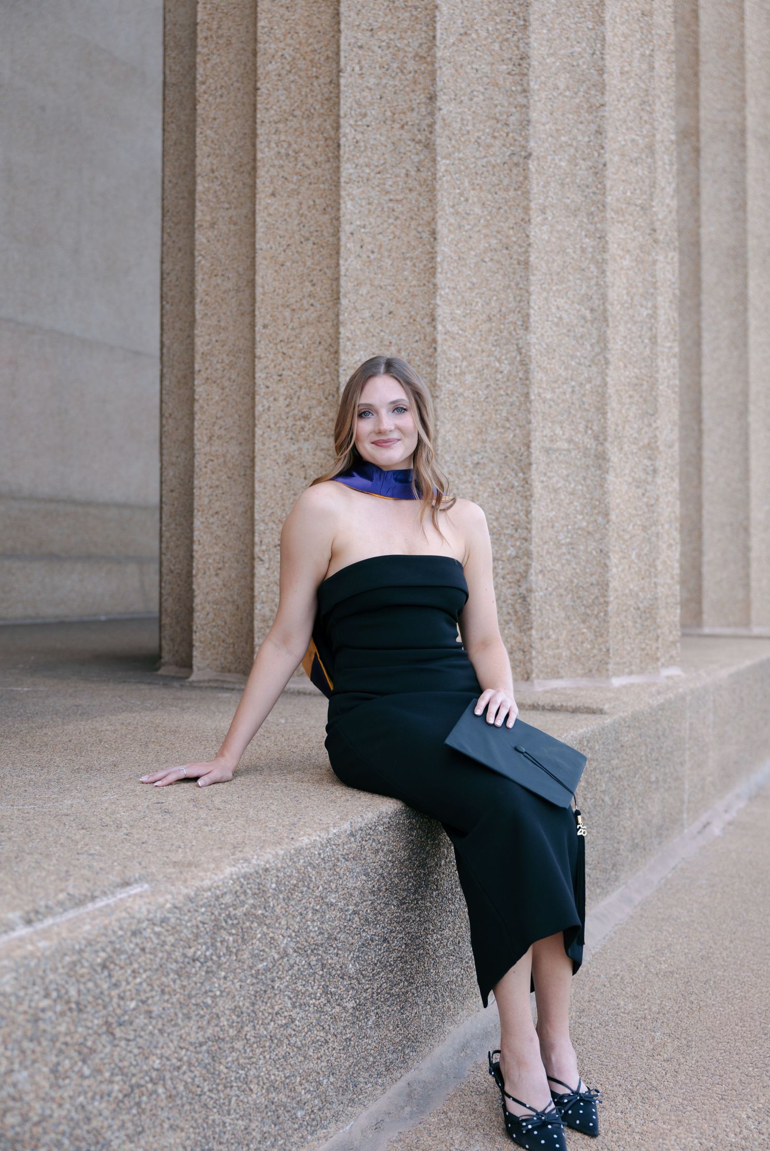 Woman in black strapless dress with purple scarf sits on stone ledge beside tall columns.