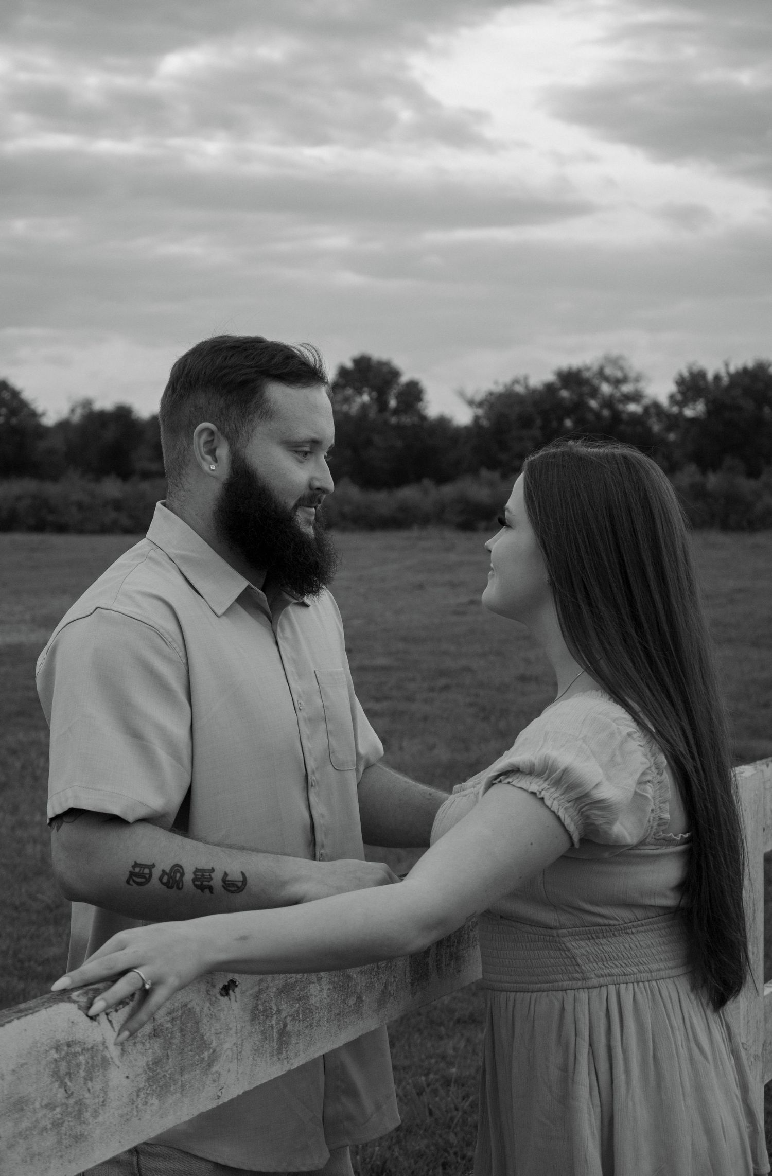 A romantic black and white portrait of a couple embracing by a wooden fence in a rural field.