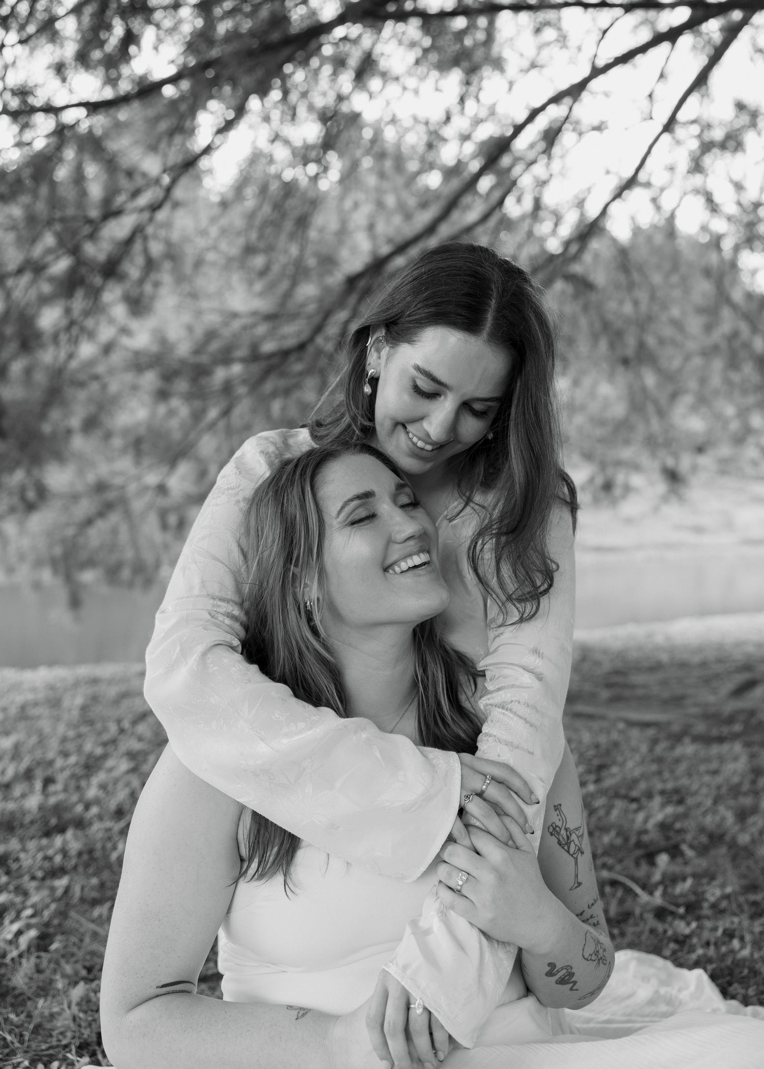 Two women share a joyful embrace outdoors in a black and white photo near a serene lake.