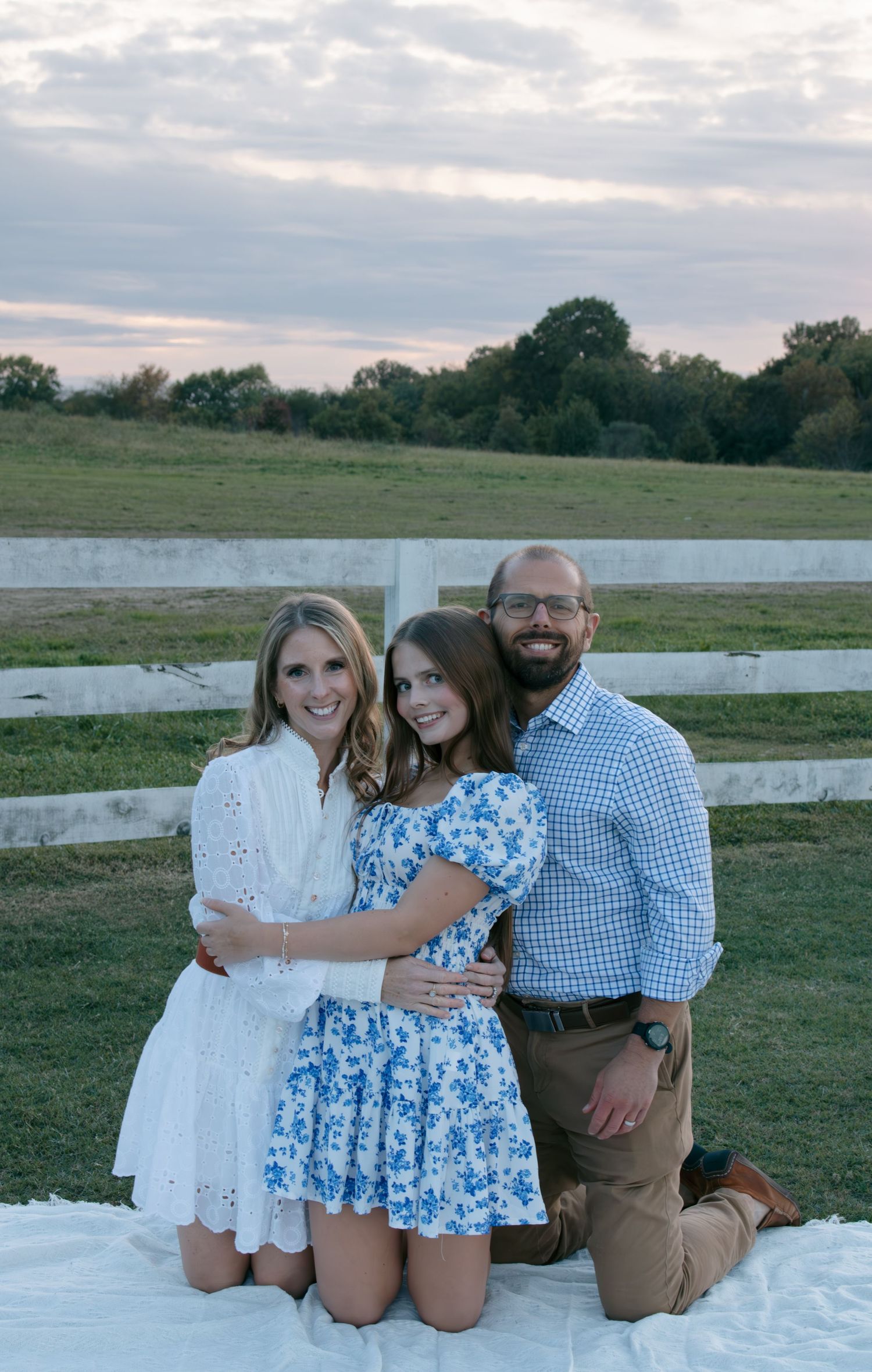 A family poses together on a white fence in a pastoral field setting at dusk.