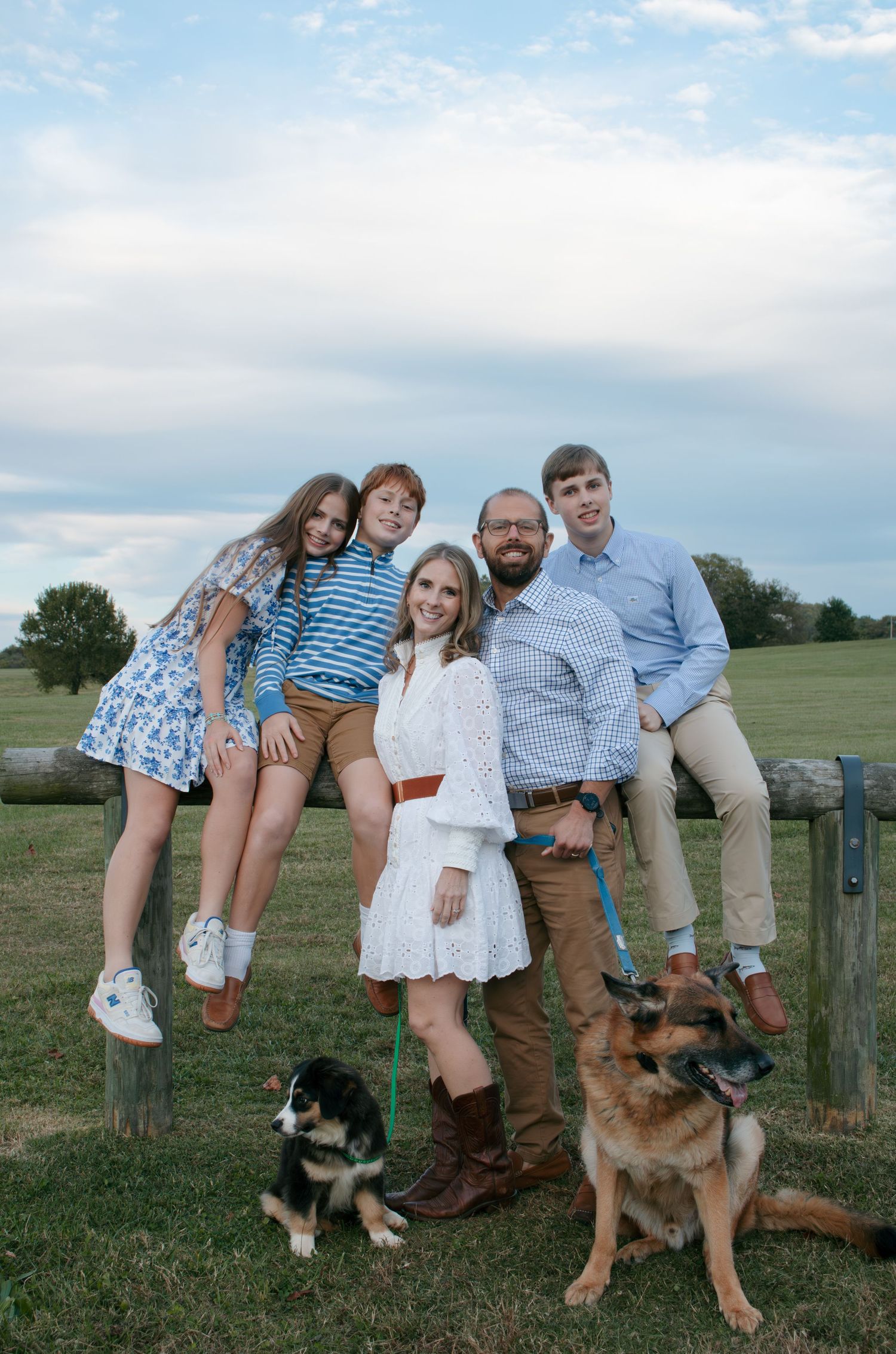 A family poses together on a wooden fence in a grassy field with their German Shepherd dog on a sunny day.