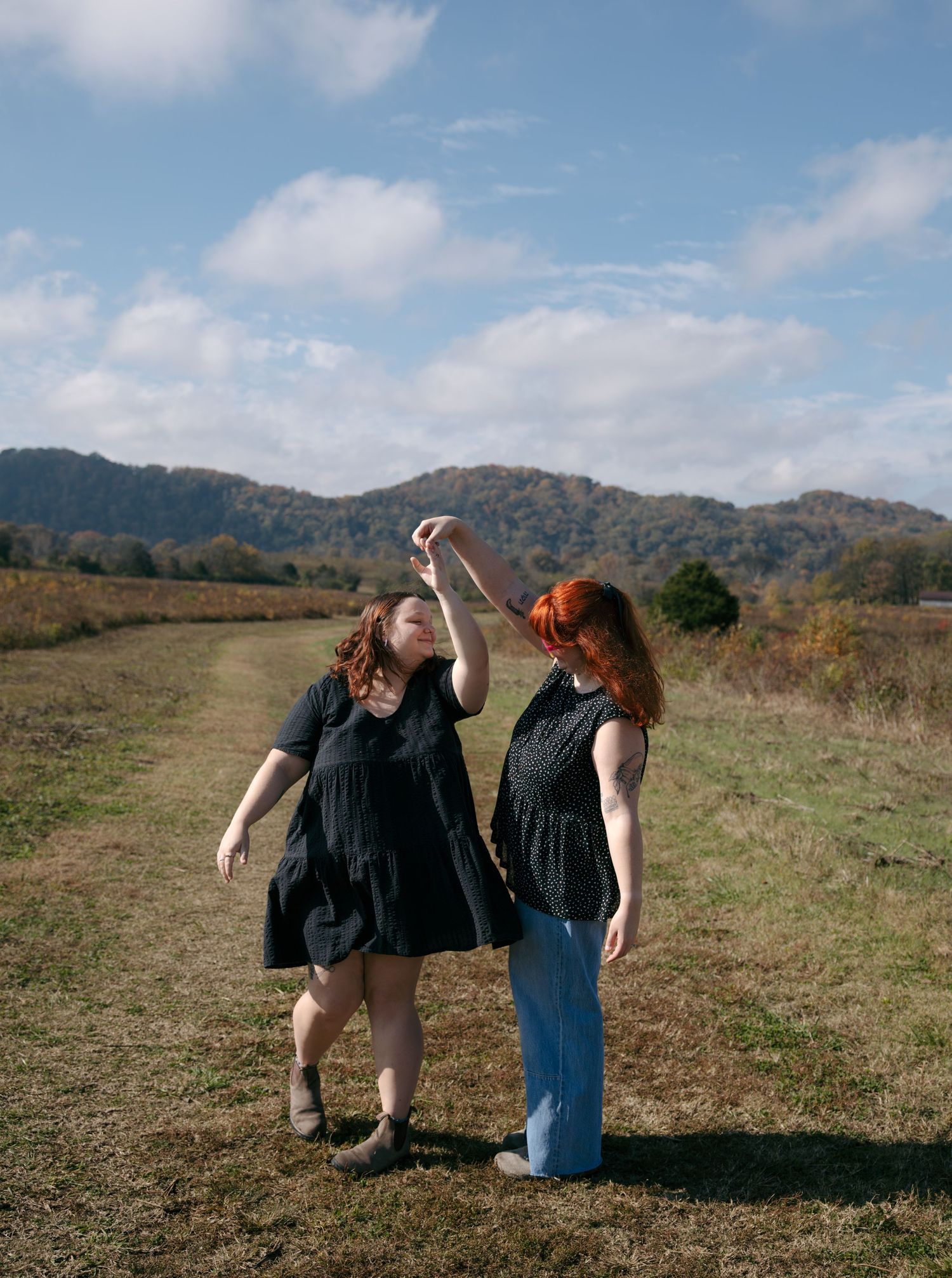 Two people in casual attire walk together on a grassy hilltop path with mountains and blue sky in the background.