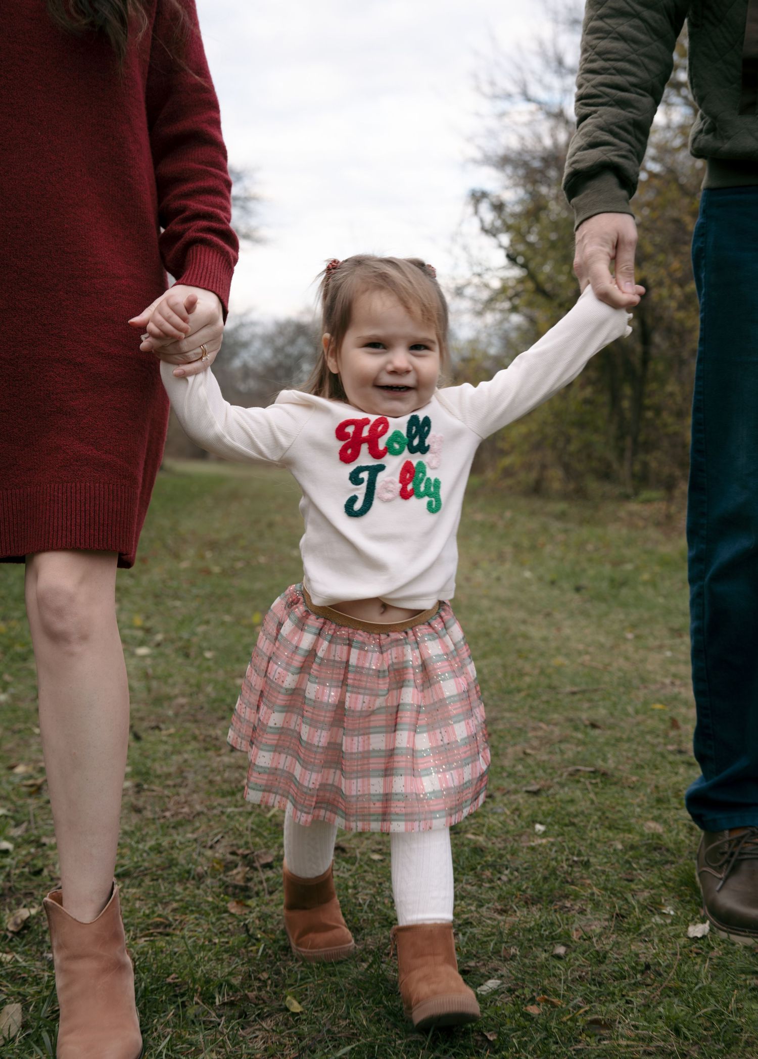 A small child in a plaid skirt and white top holds hands with adults while joyfully jumping in a grassy outdoor setting.