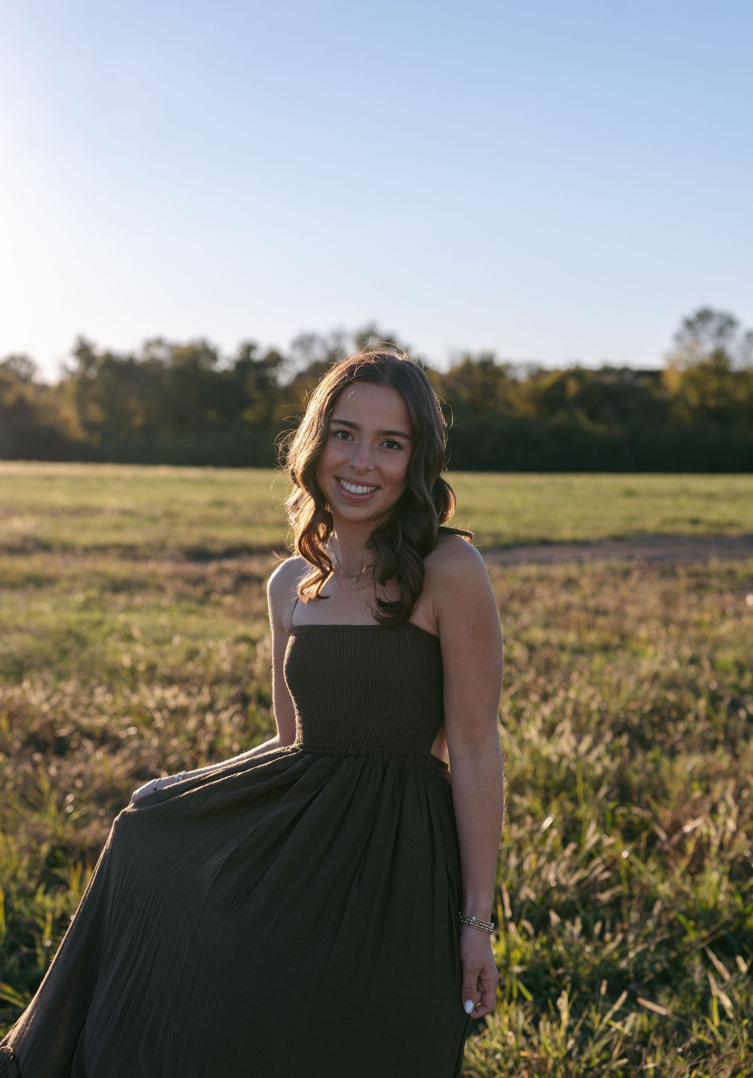 Smiling woman with long wavy hair in a dark green strapless maxi dress standing in a golden grassy field at sunset with trees behind.
