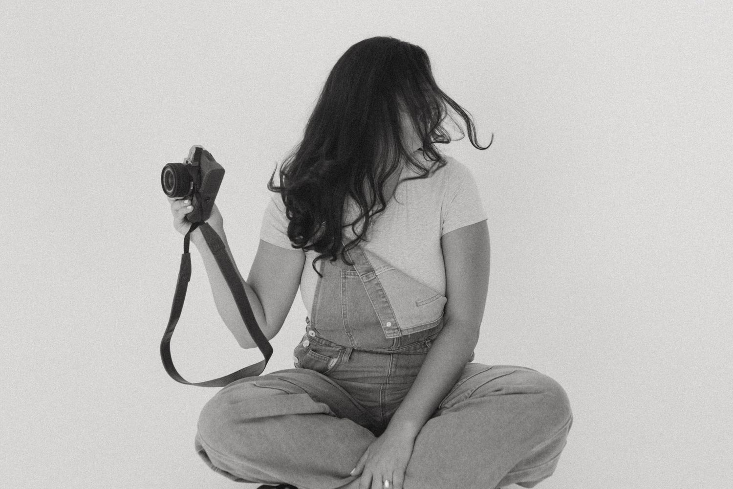 Photographer in casual denim outfit sits cross-legged on white background holding professional camera.