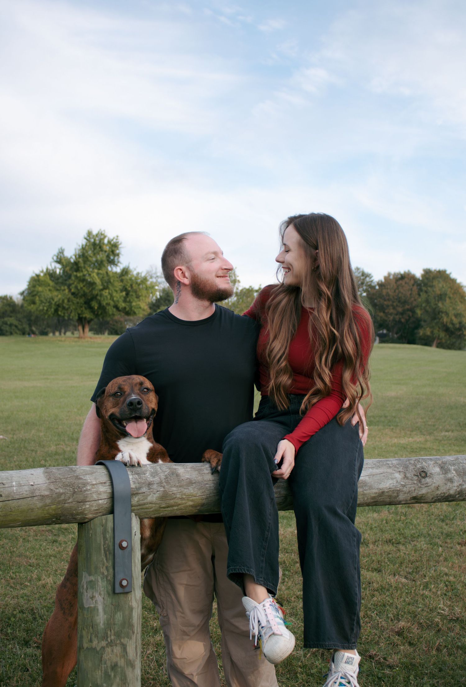 Couple and their dog relaxing on wooden fence in grassy field.