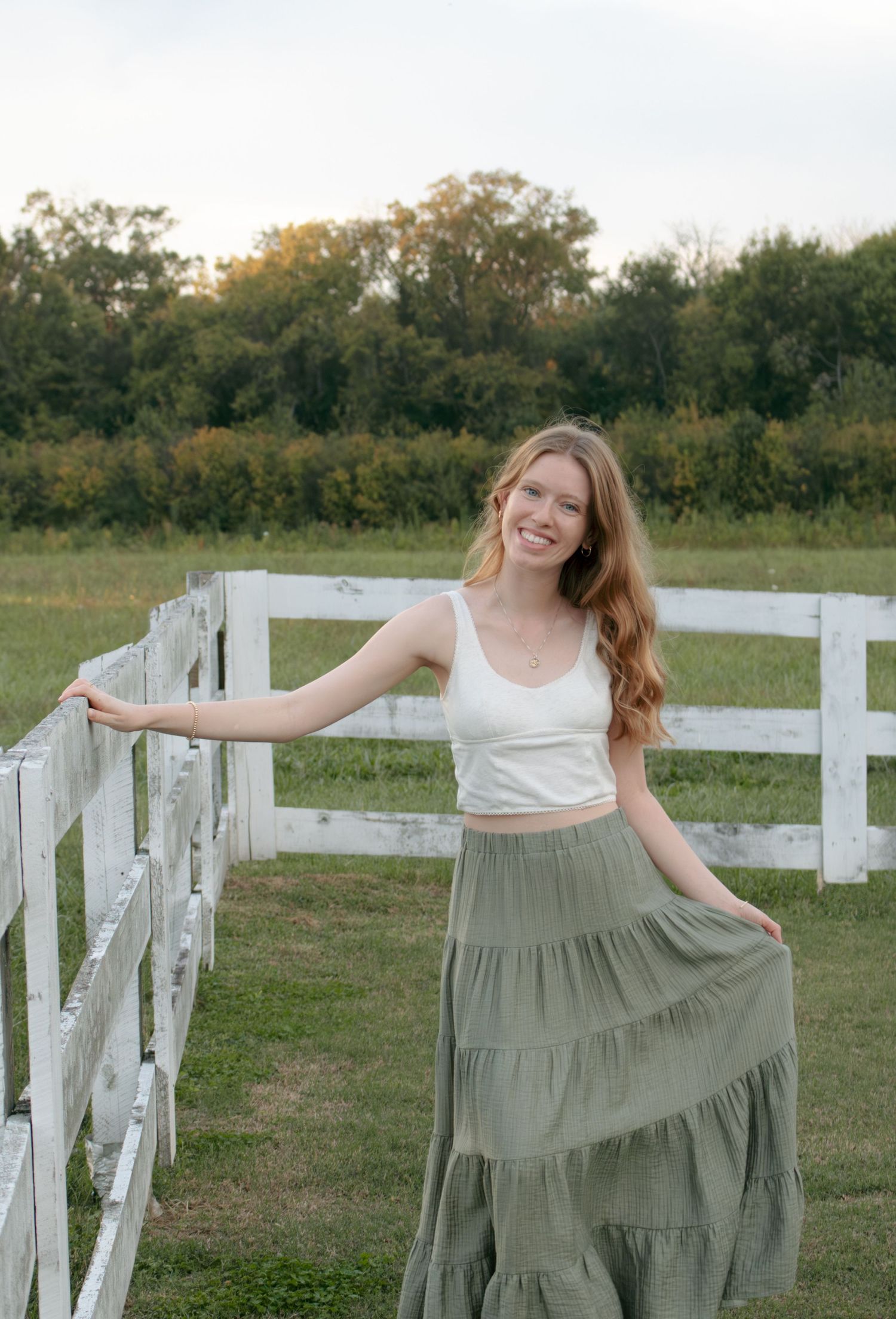 Person twirling in flowy sage green dress by white fence in countryside setting.