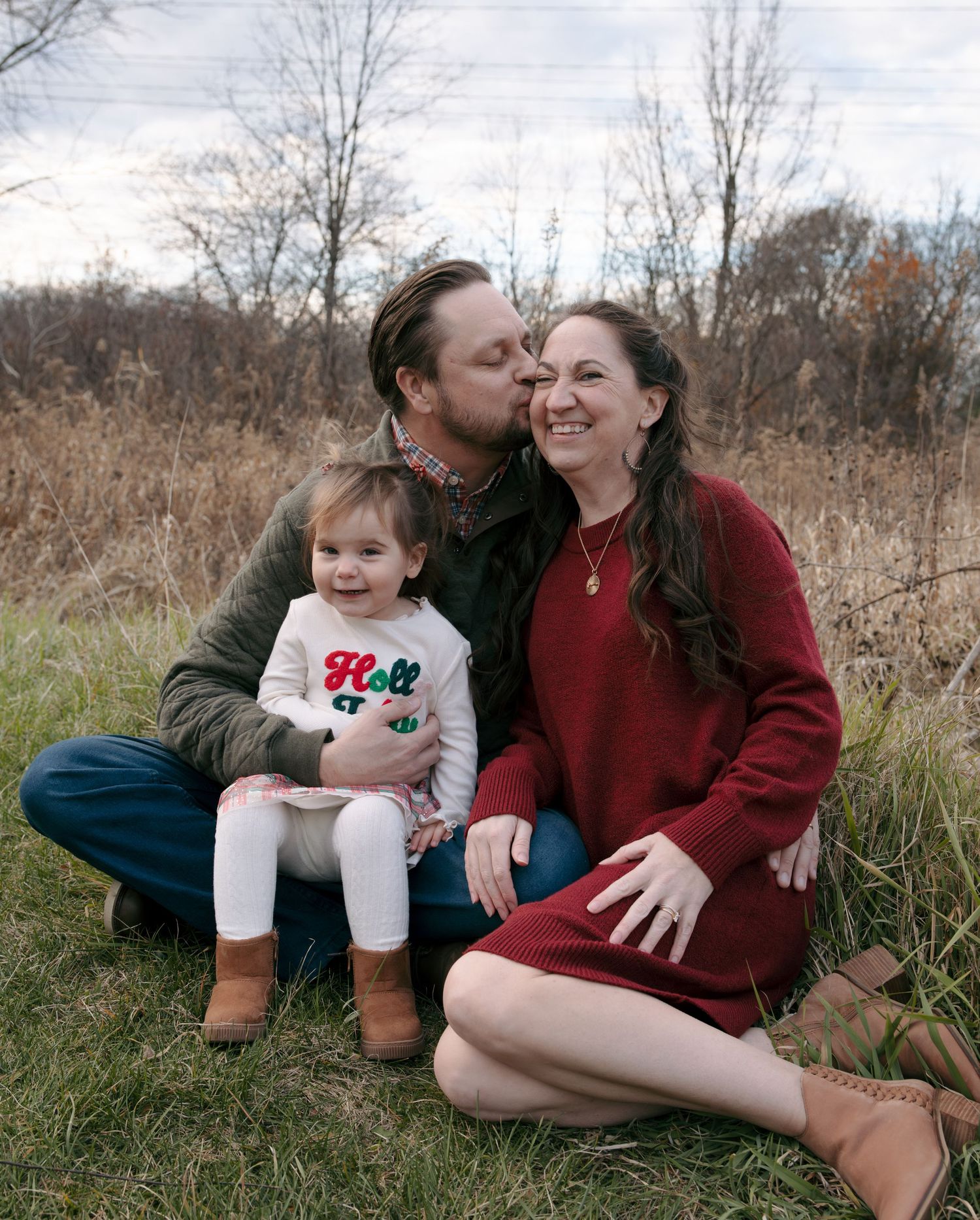 A family sits together in a grassy field during autumn, wearing casual clothing and sharing an affectionate moment.