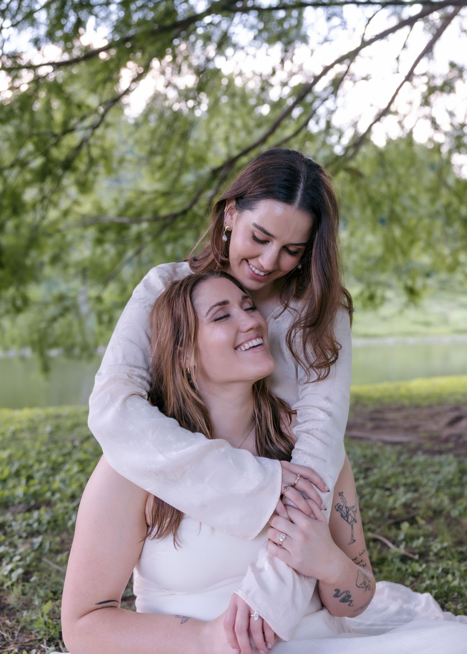 Two women in white dresses share a tender embrace outdoors near a tree by a lake.