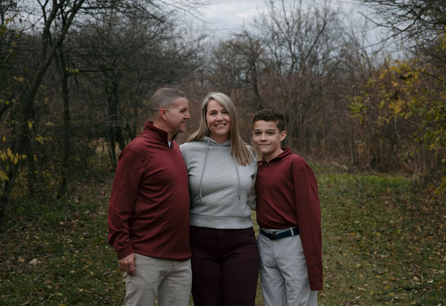 A family wearing burgundy and gray sweaters poses together for outdoor autumn photos in a wooded setting.