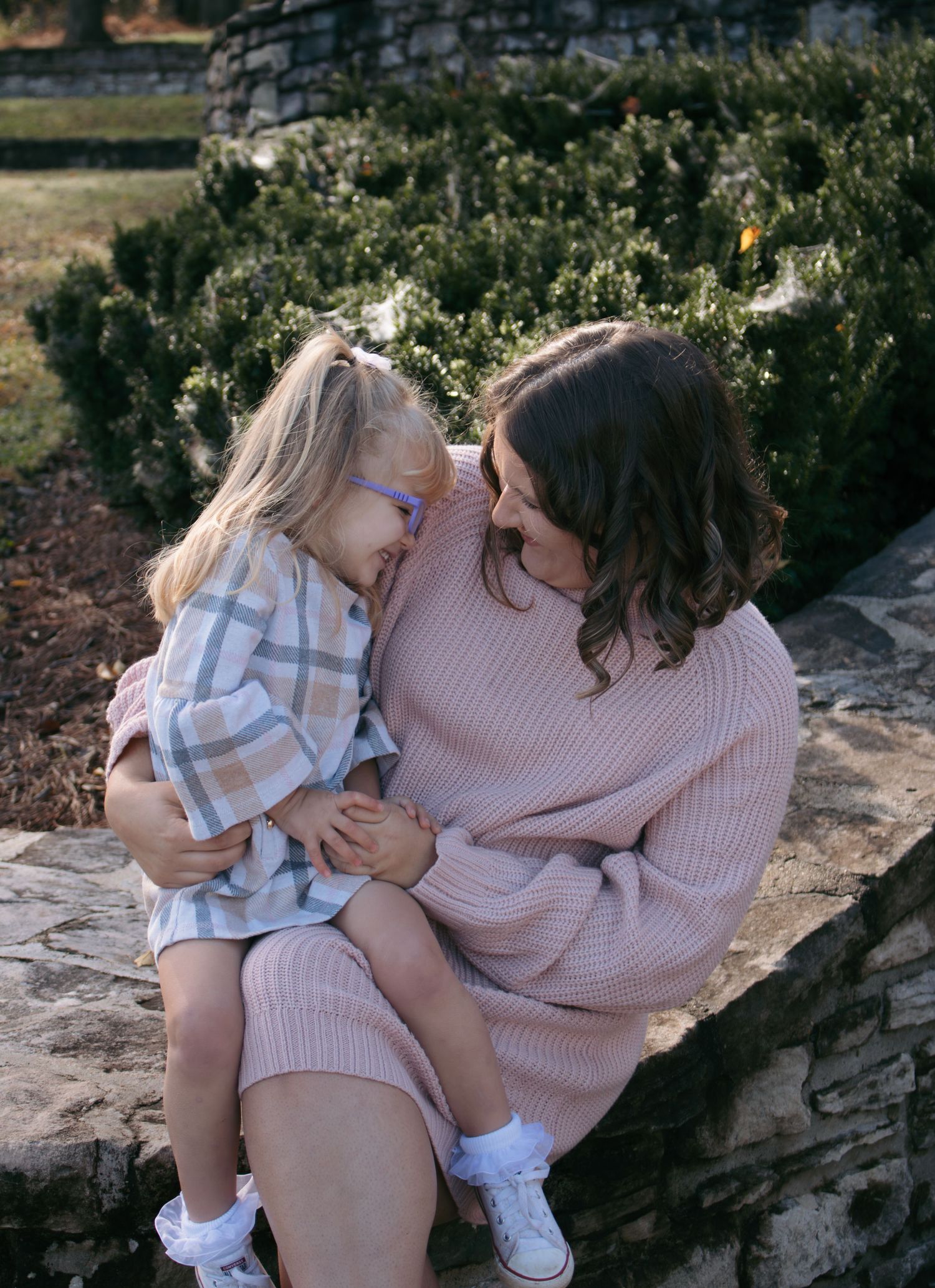 Two people in coordinating pink and plaid outfits sit together on rocky terrain during a sunny outdoor photoshoot.