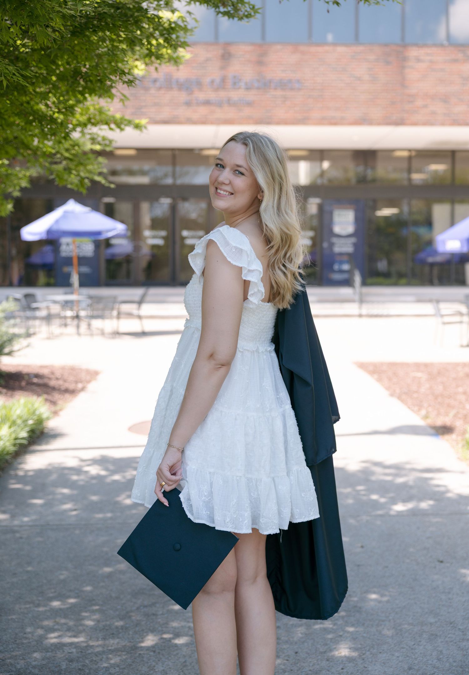 Young woman in white dress holding graduation cap and gown, smiling outdoors near a College of Business building.