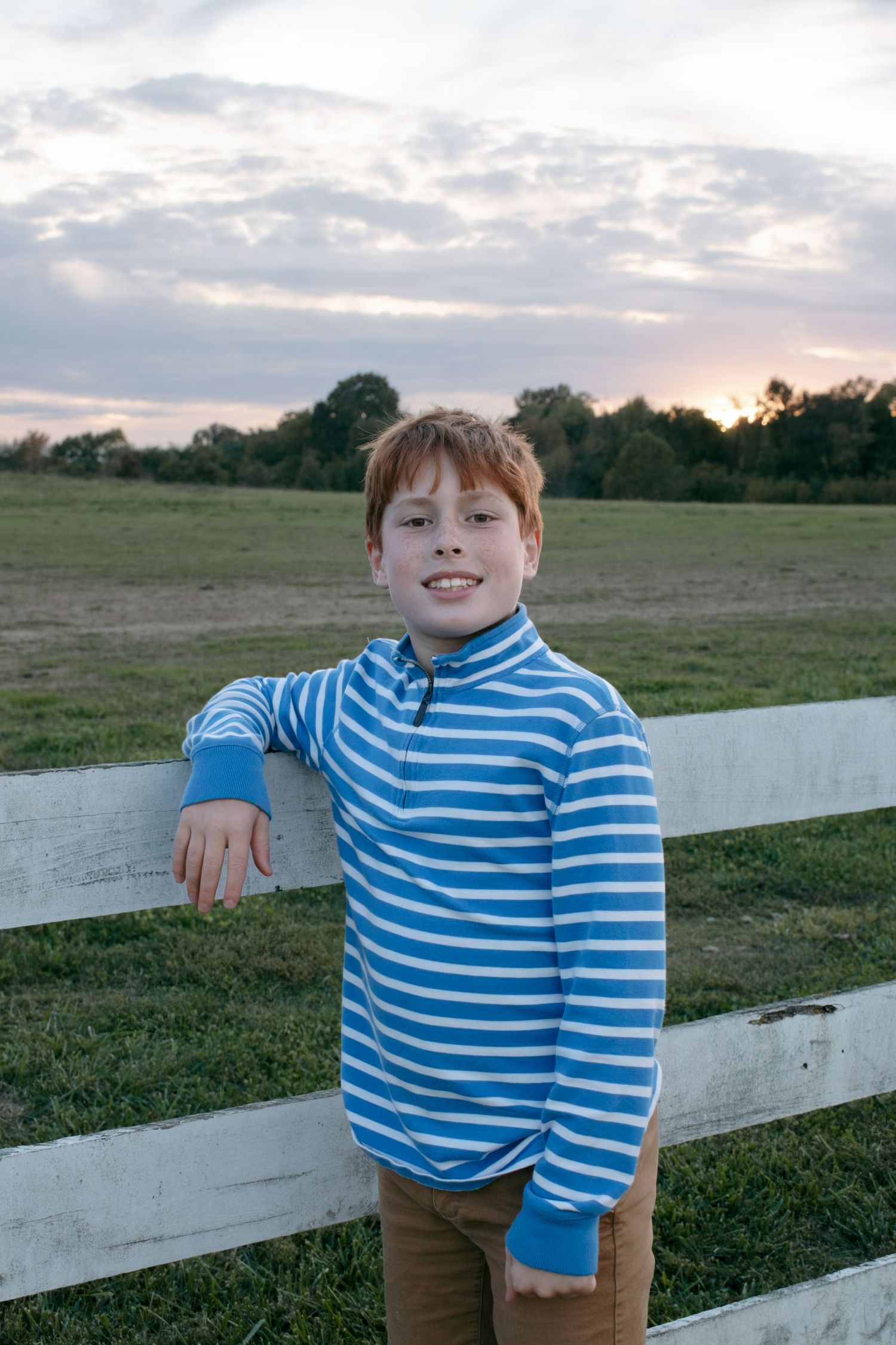 A child in a blue striped shirt leans against a white fence at sunset in a grassy field.