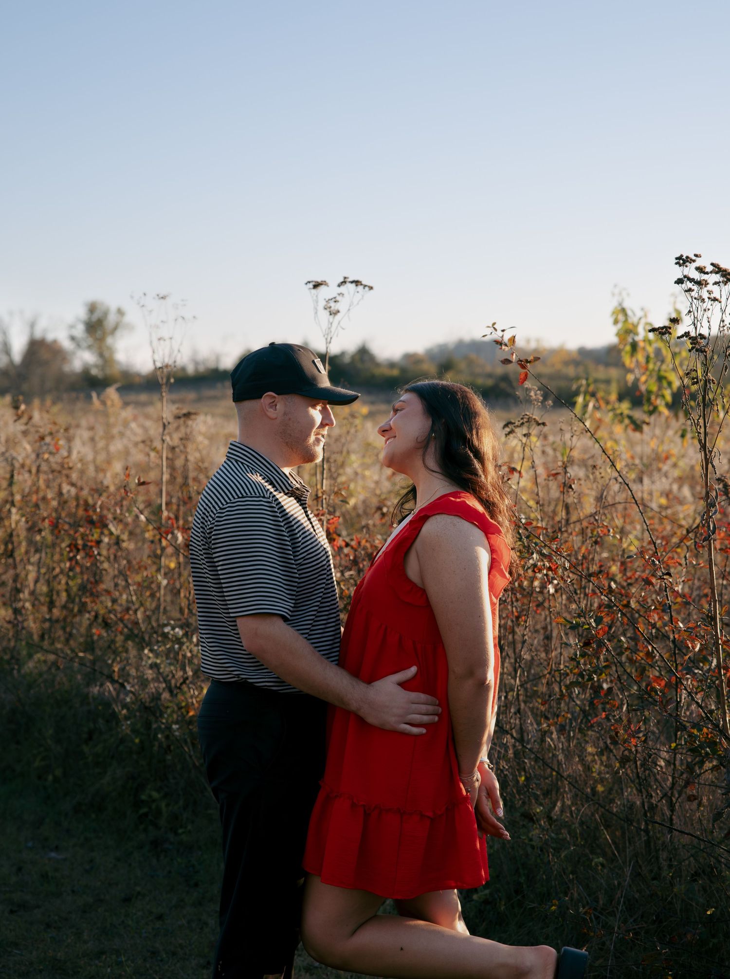 A couple shares an intimate moment in a golden field during sunset, with wildflowers and tall grass surrounding them.
