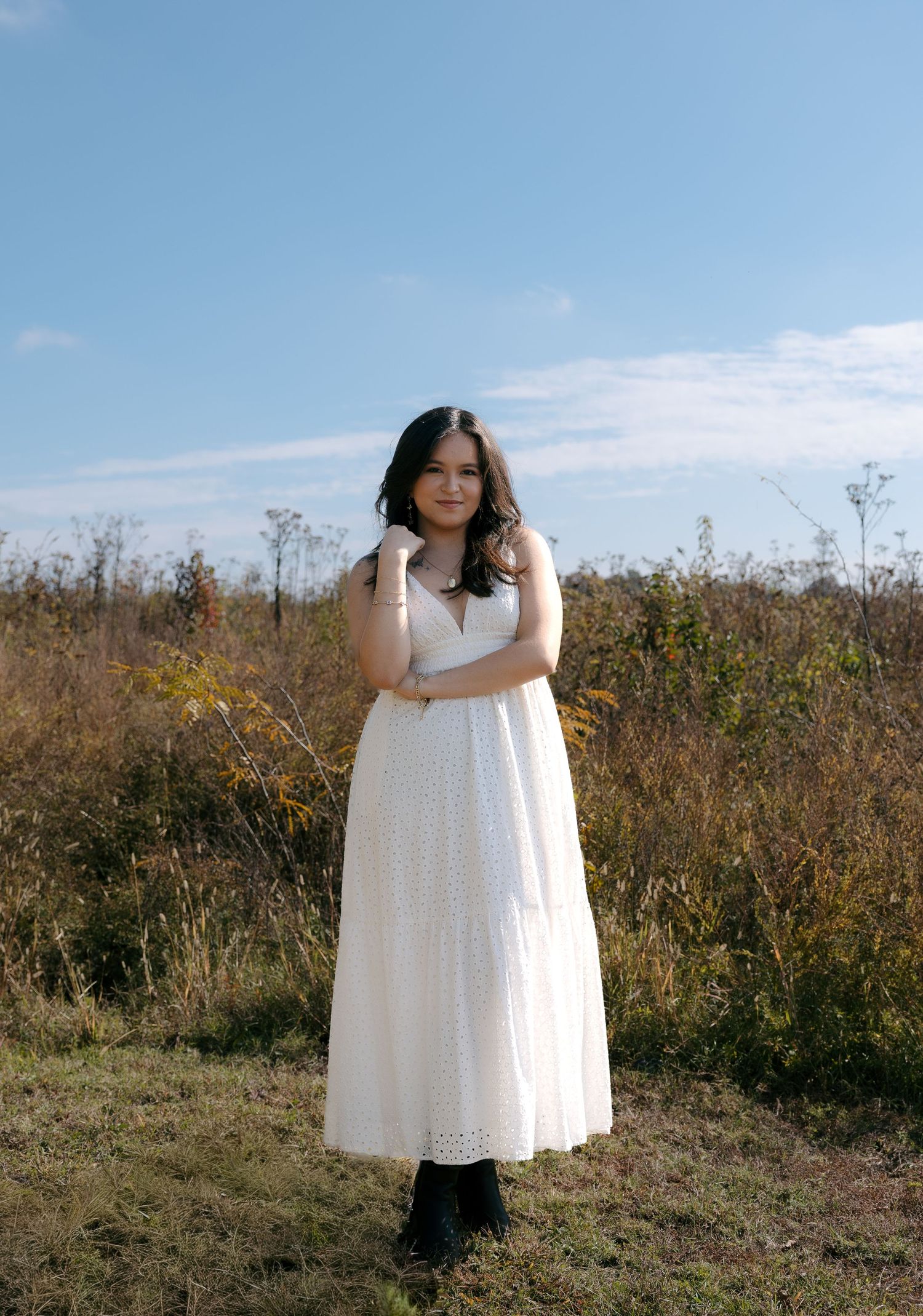 A person in a flowing white dress poses gracefully in a natural field setting against a bright blue sky.