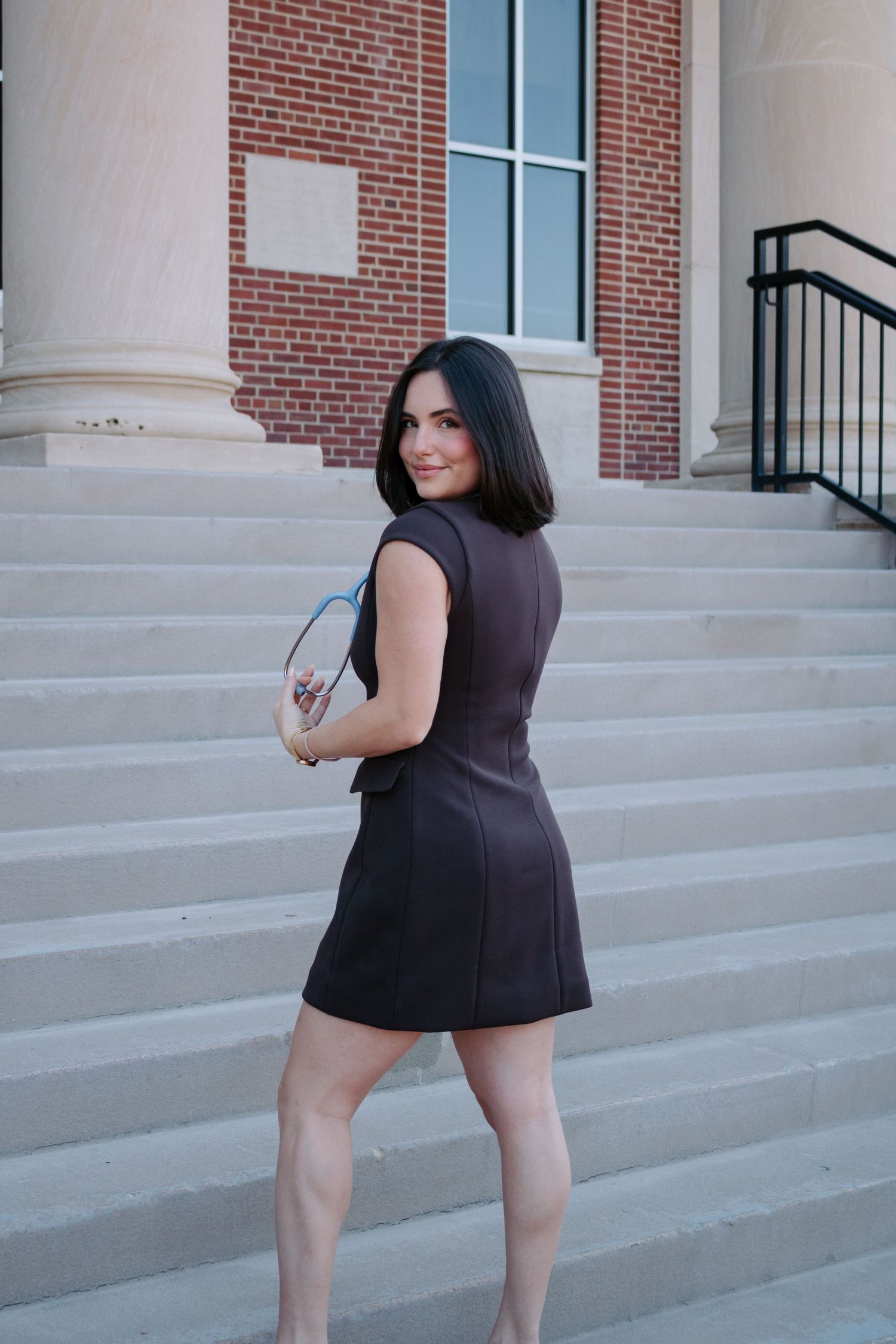 Woman in a dark dress poses on stone steps in front of a brick building with white columns.