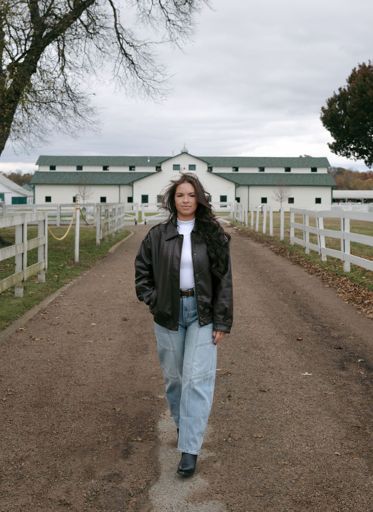A figure in a black jacket and light jeans walks along a dirt path bordered by white wooden fences on an overcast day.