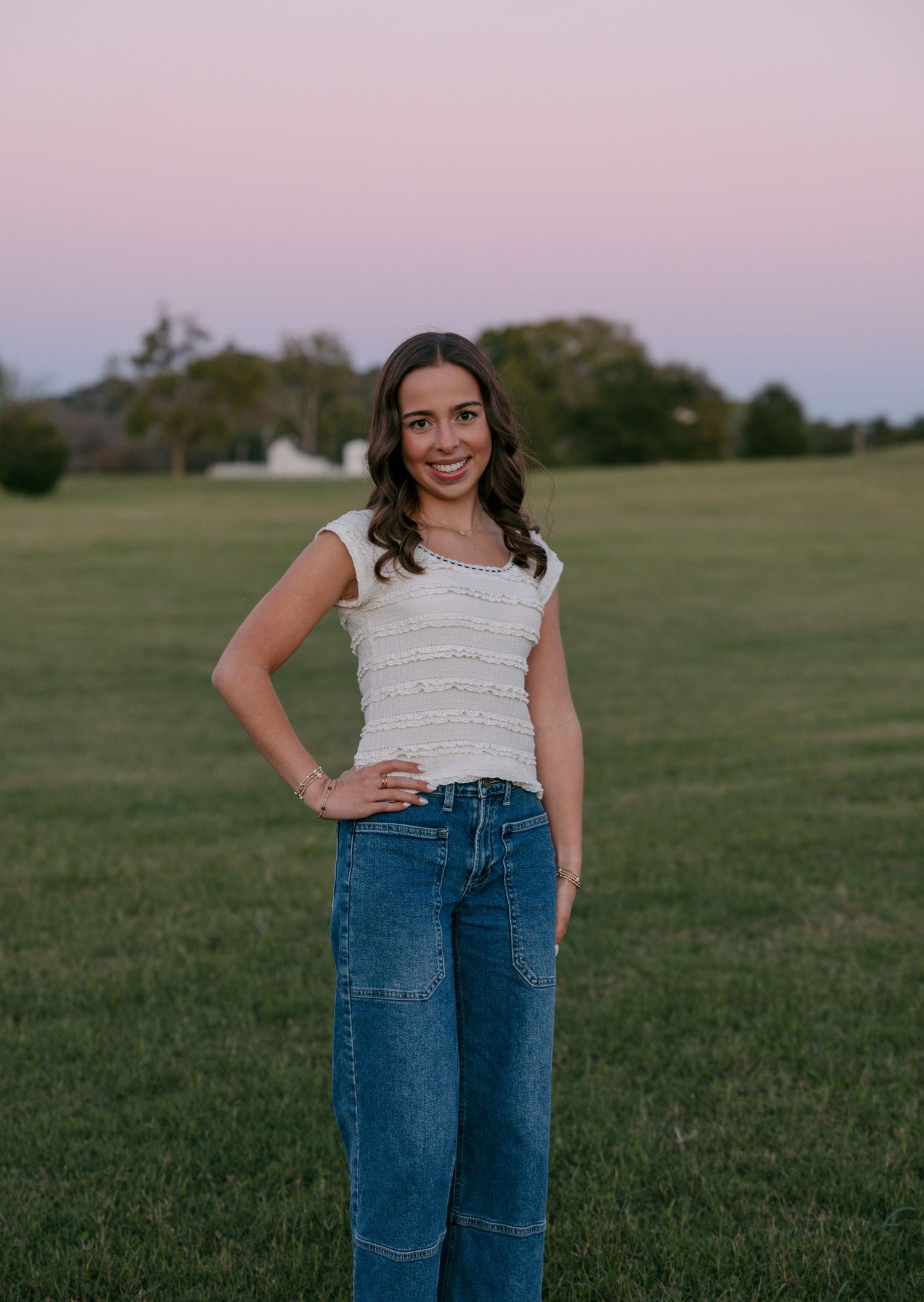Smiling young woman with wavy brown hair, white knit top and wide-leg jeans, standing with hand on hip in a grassy field at dusk.