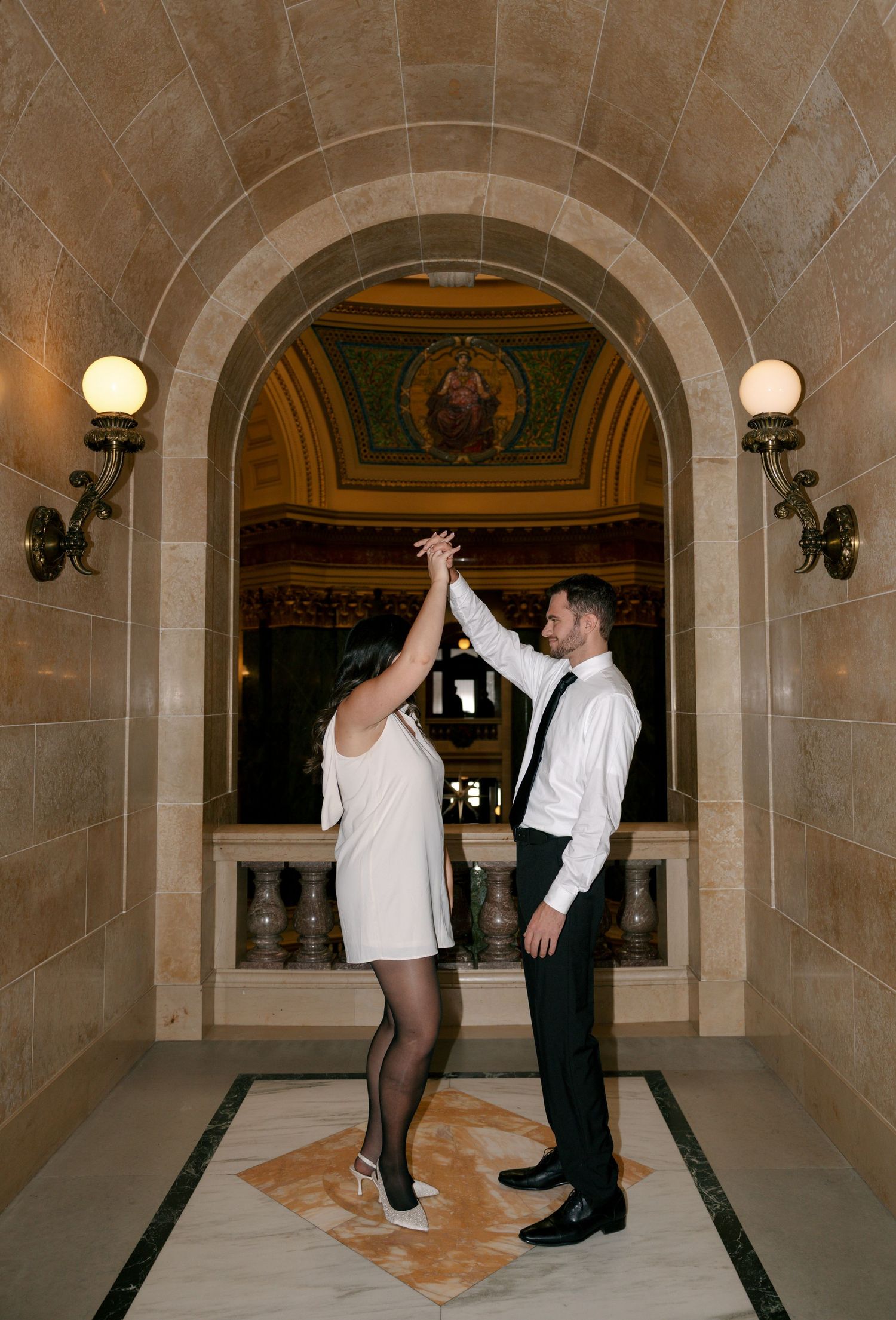 A couple performs a series of dance moves in an elegant marble hallway with arched ceilings and wall sconces.