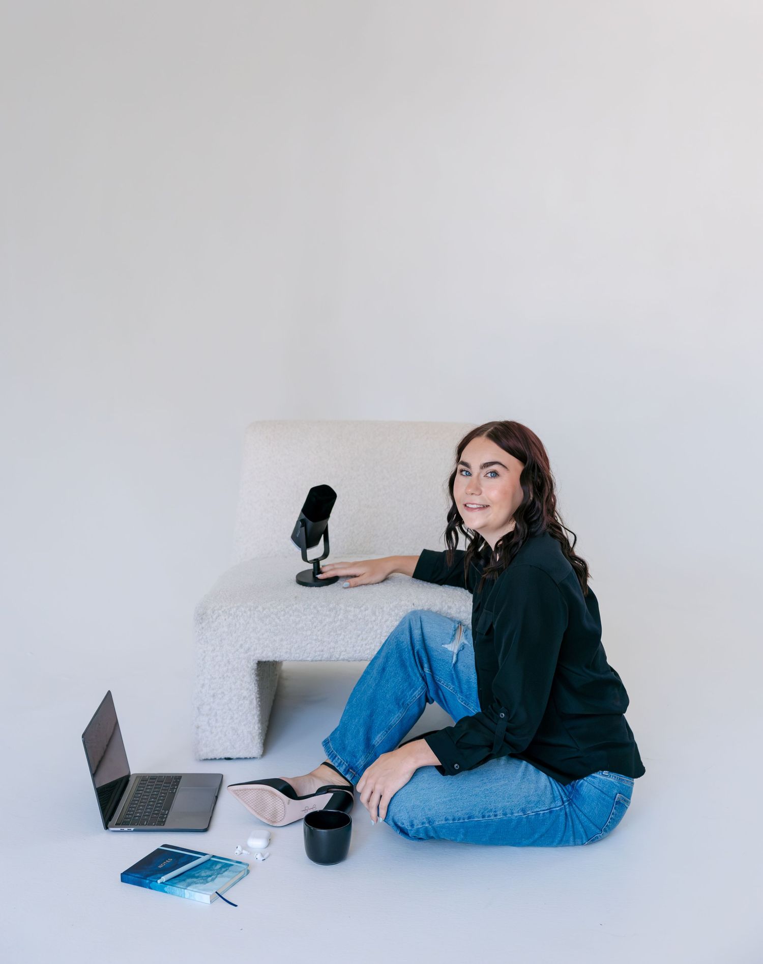 Woman sitting on floor beside a microphone and laptop, smiling at camera in a bright white studio setting.