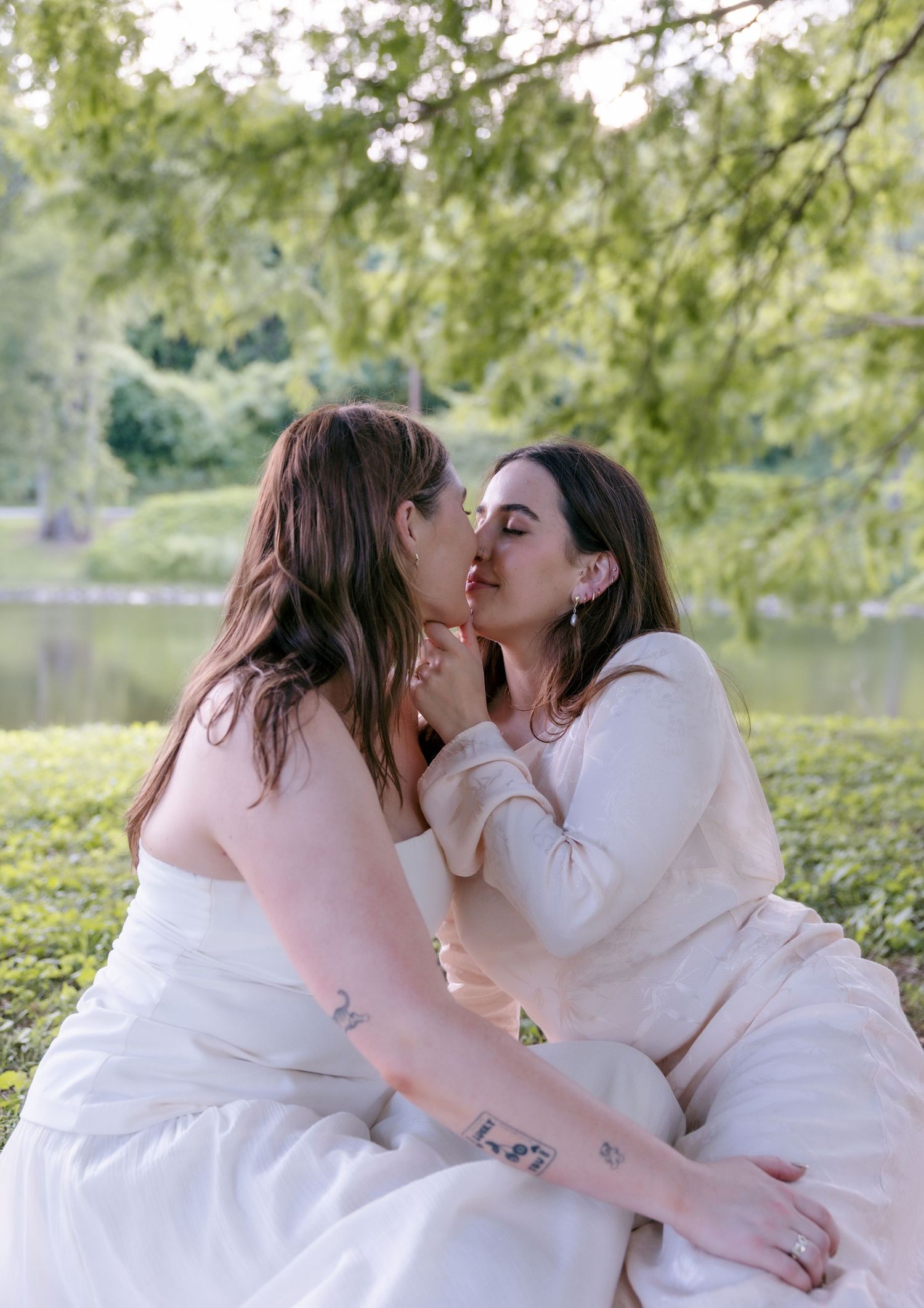 Two brides sharing a romantic kiss outdoors near a pond, surrounded by lush green trees on their wedding day.