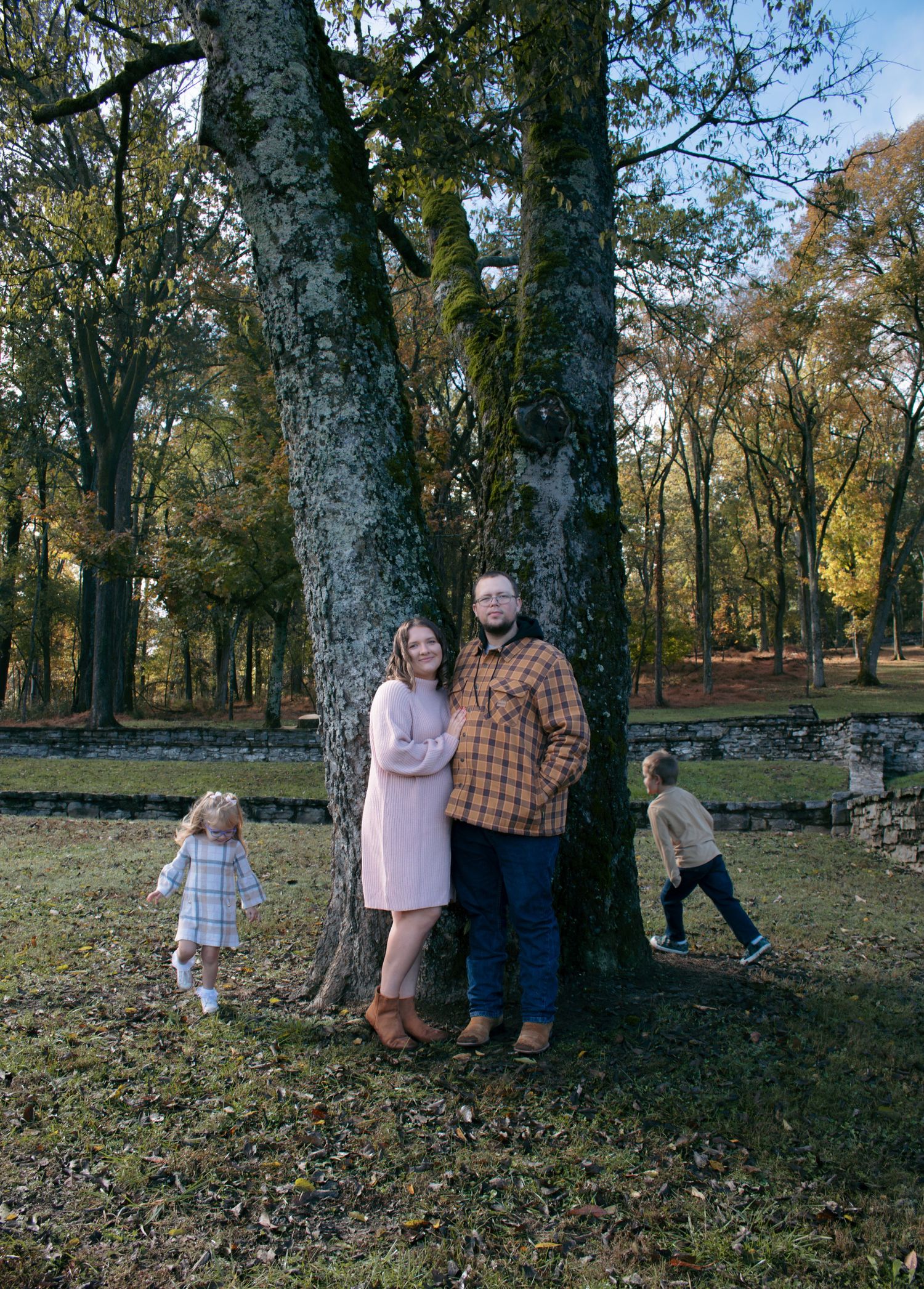 A family poses for autumn photos among tall trees in a park with fallen leaves scattered on the ground.