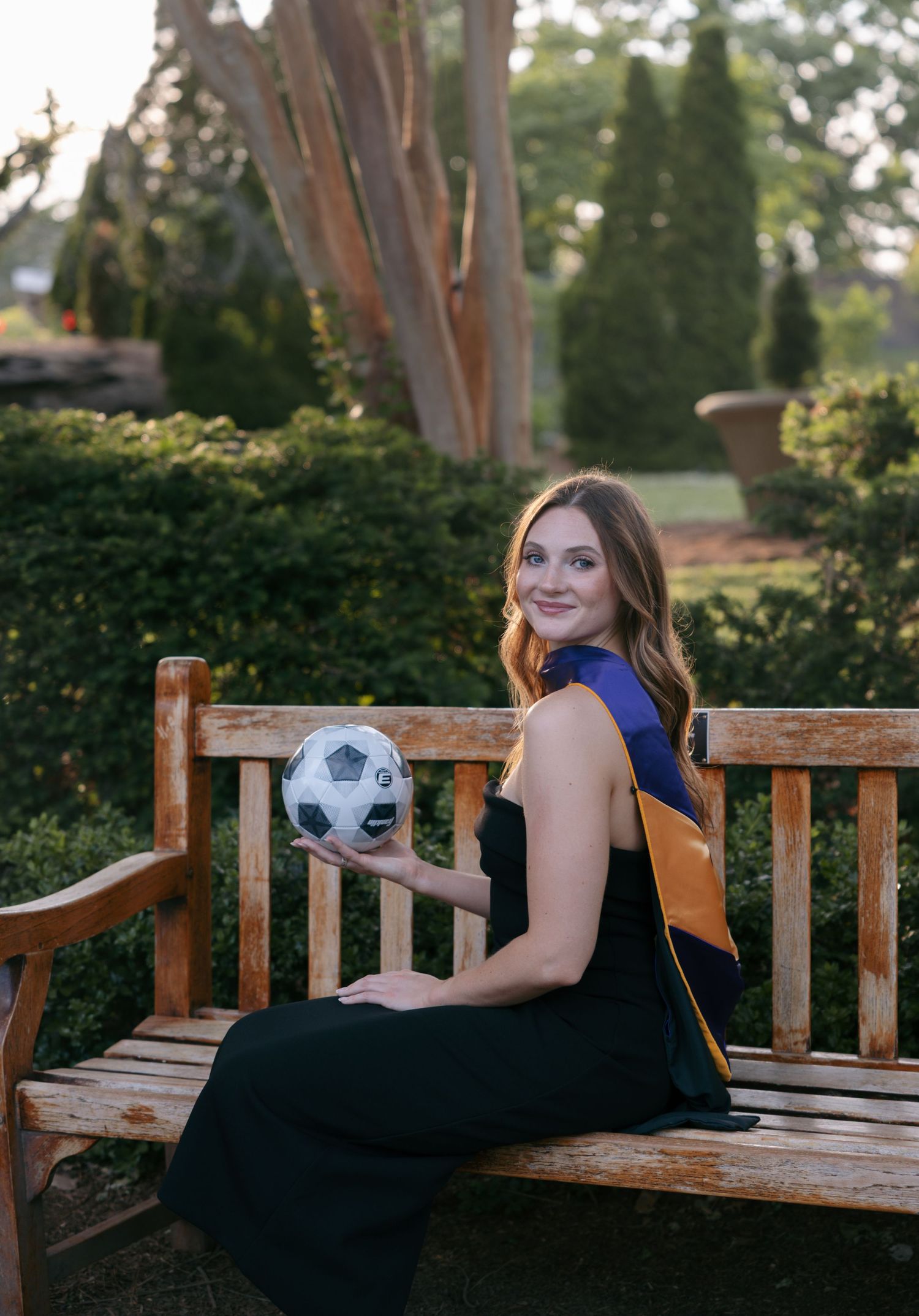 A smiling young woman in graduation regalia holds a soccer ball while sitting on a wooden bench in a lush garden.