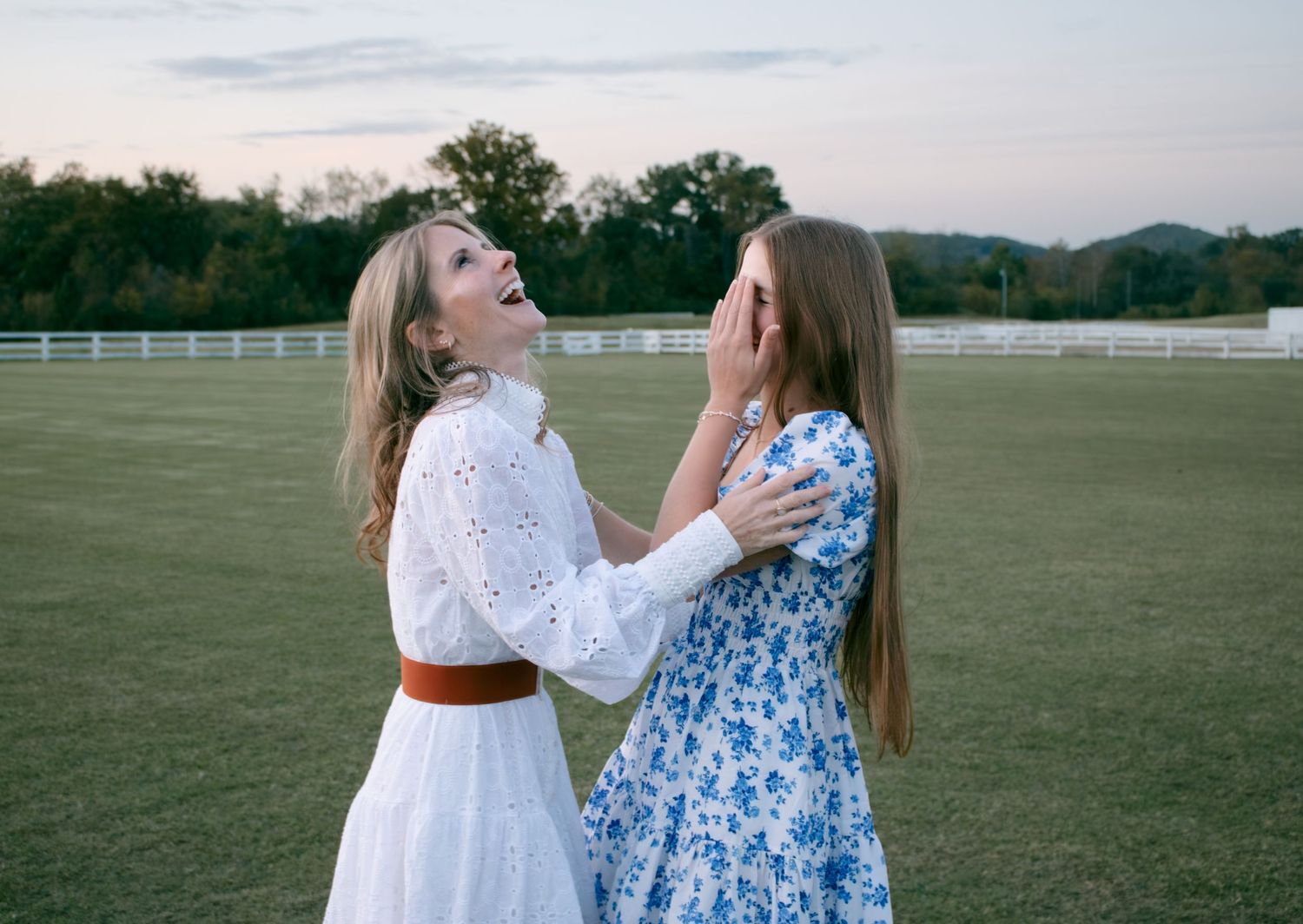 Two people in white and blue dresses dance together in a grassy field at sunset.