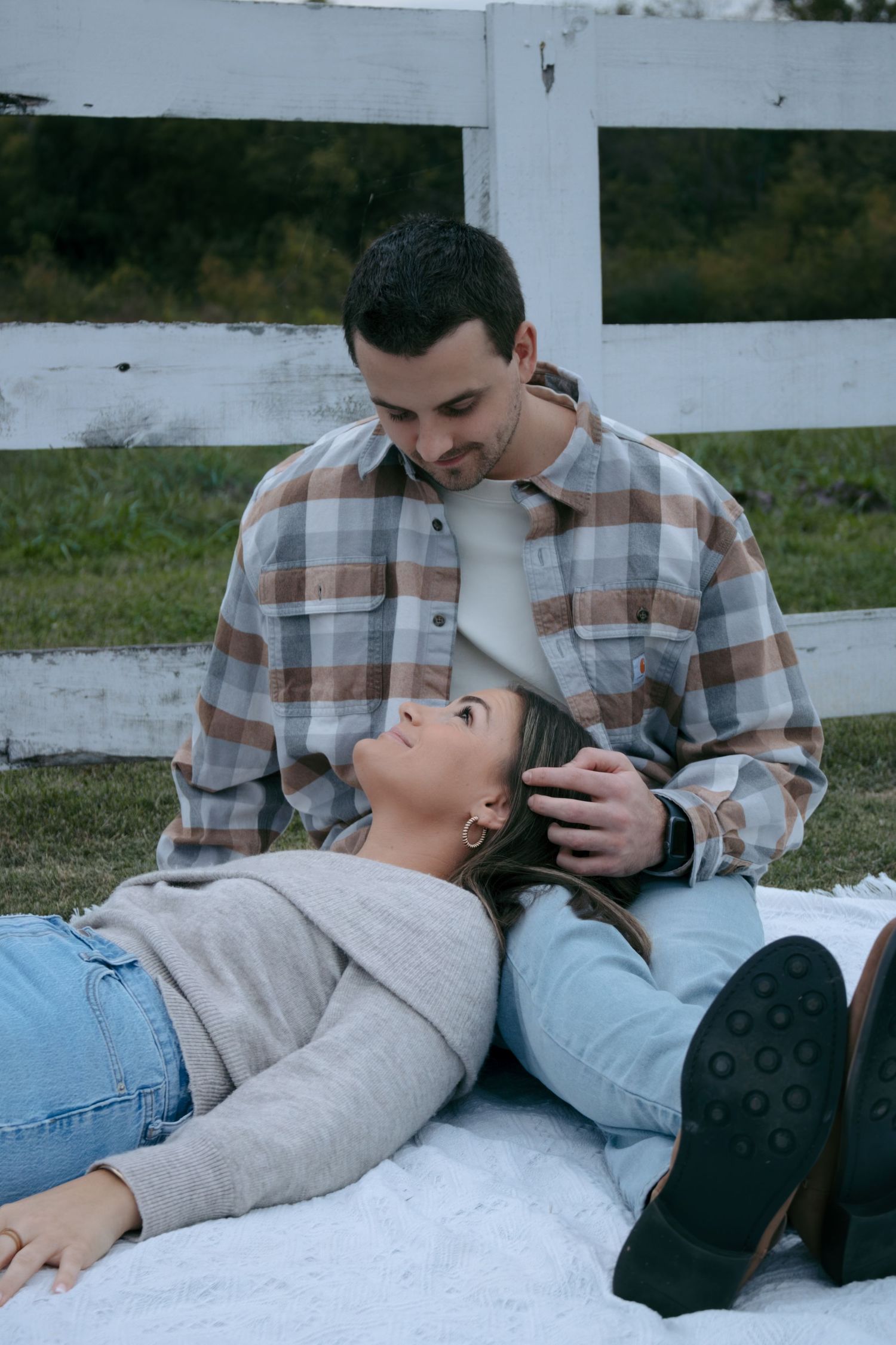 Couple sharing an intimate moment while lying on a blanket outdoors near a white fence.