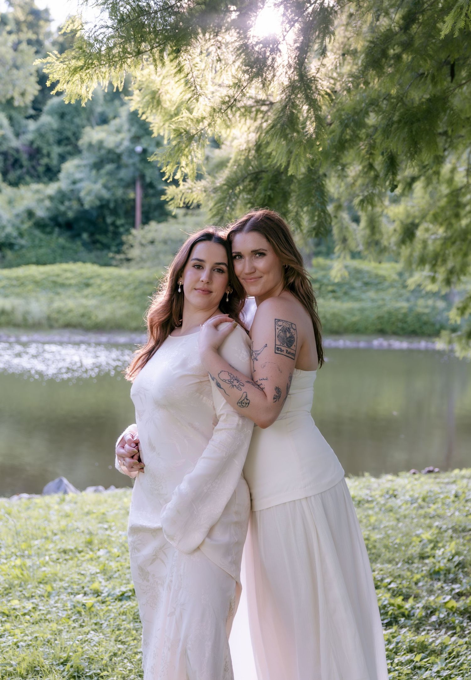 Two brides in white gowns embrace outdoors near a pond, one with tattoos, surrounded by lush greenery.