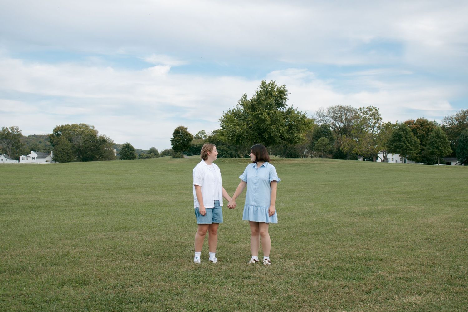 Two people in light colored clothing stand together in a vast green field on a summer day.