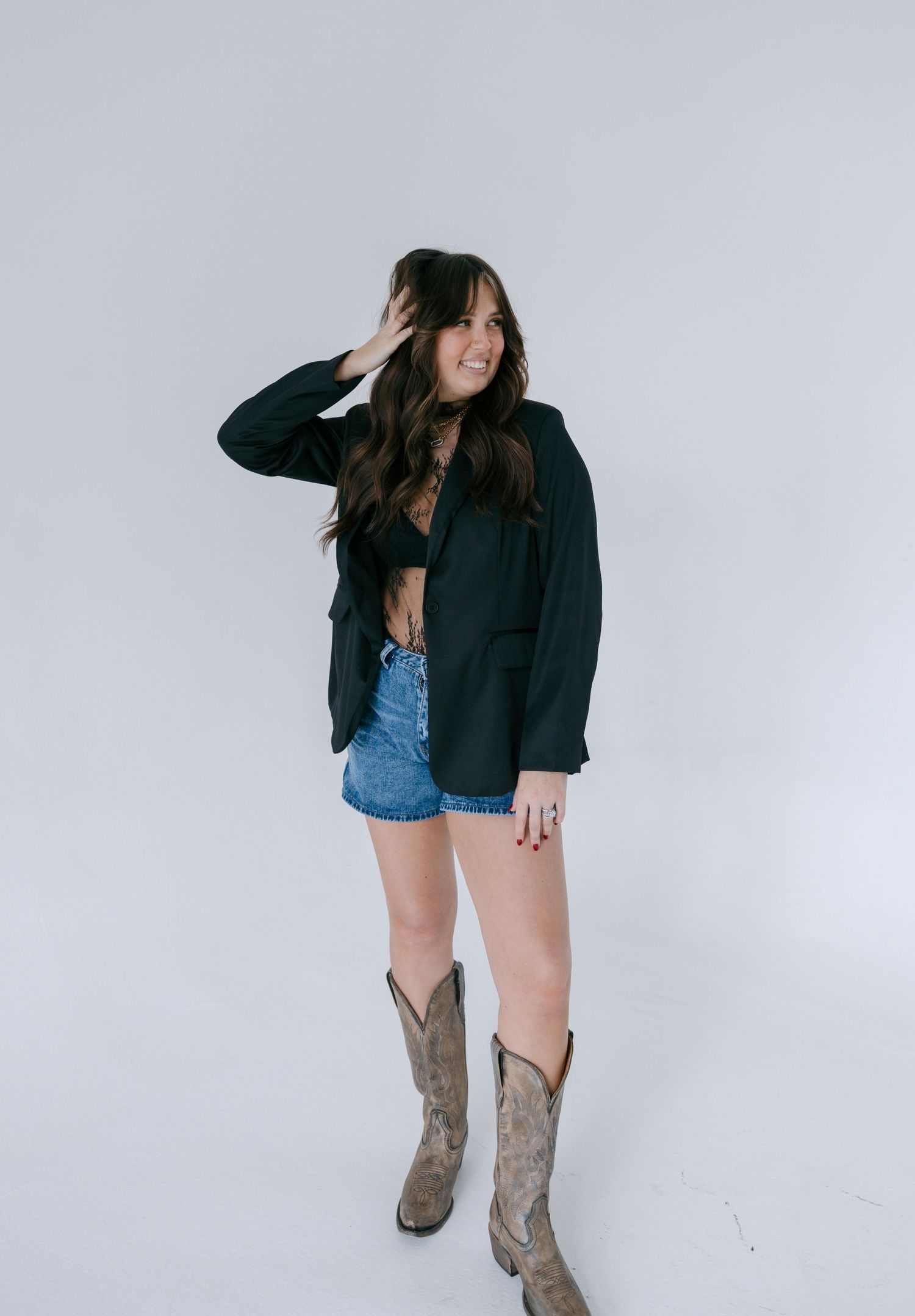 A person wearing a black blazer, denim shorts and cowboy boots poses against a white background in multiple shots.